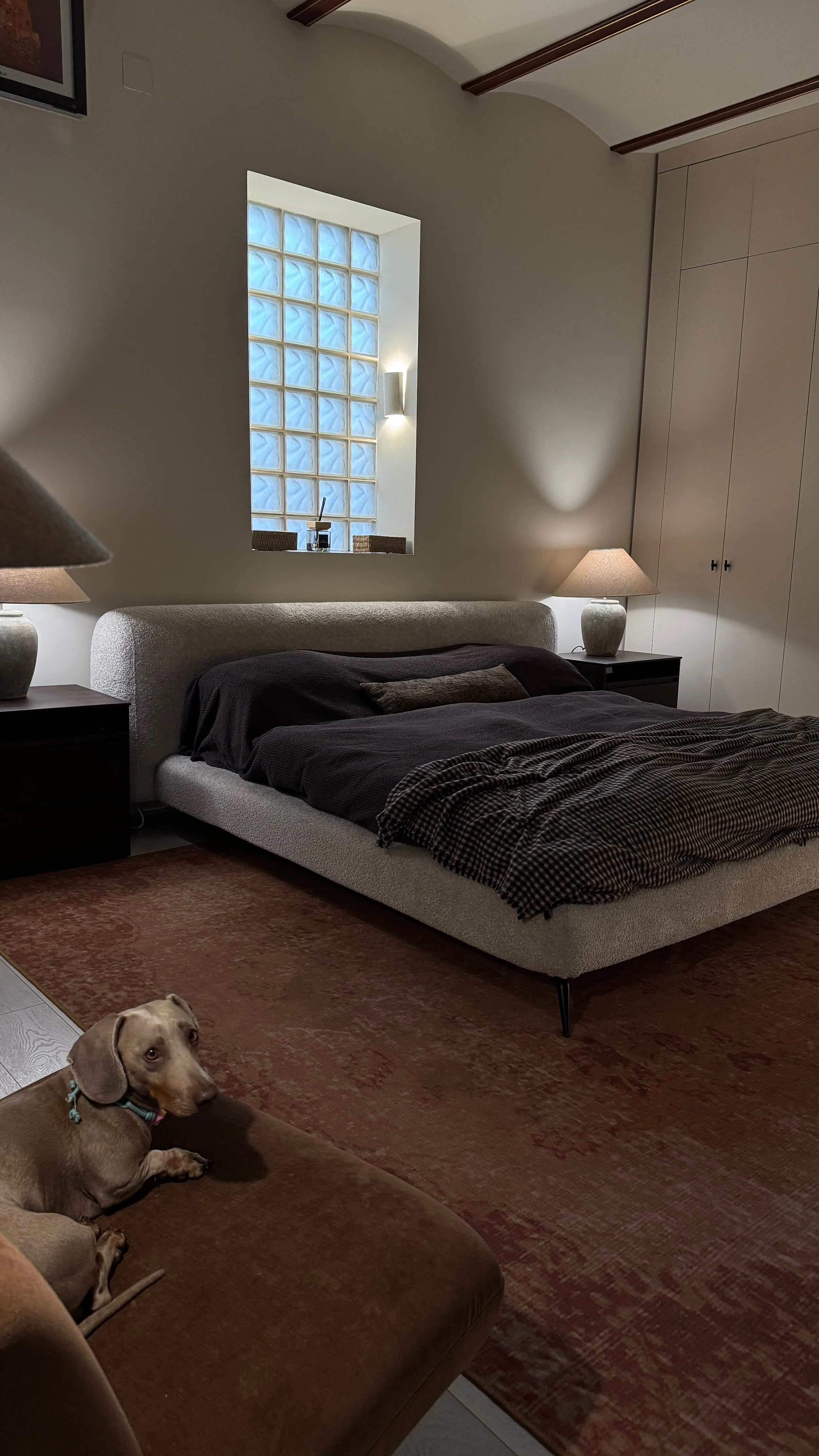 A bedroom with a low bed, two nightstands with lamps, a window made of glass blocks, a closet, and a dog lying on a brown couch.