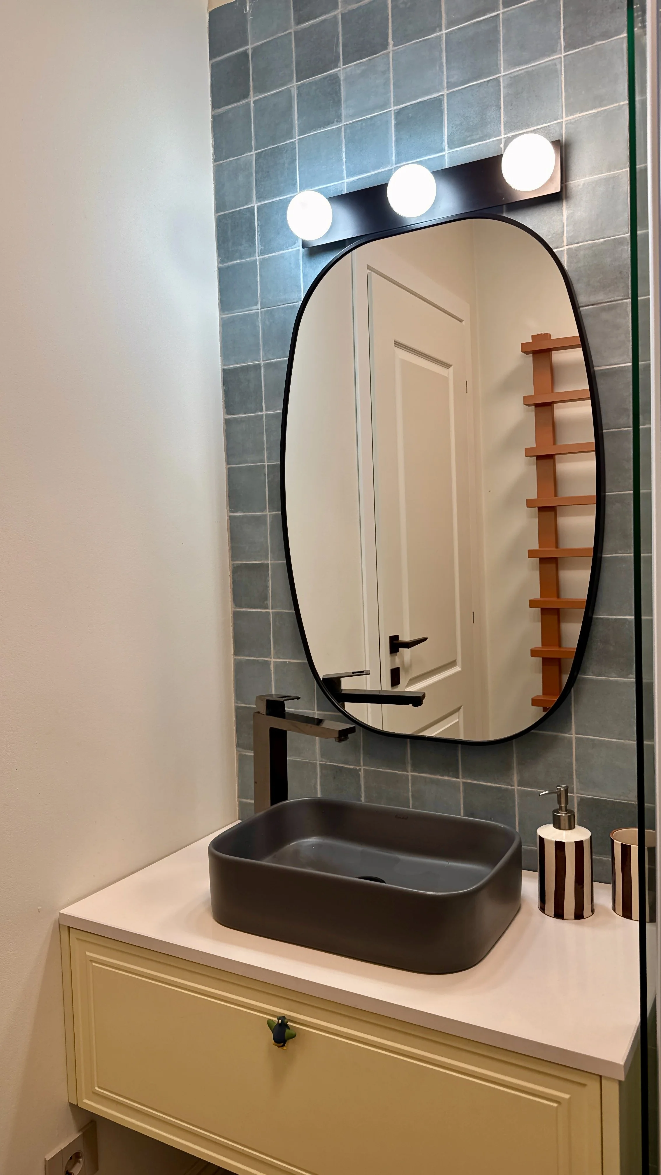 A bathroom vanity with a white countertop and a gray vessel sink. A mirror is mounted above the counter, and a light fixture with three round bulbs is above the mirror. There are two striped containers and a soap dispenser on the countertop, and a wo