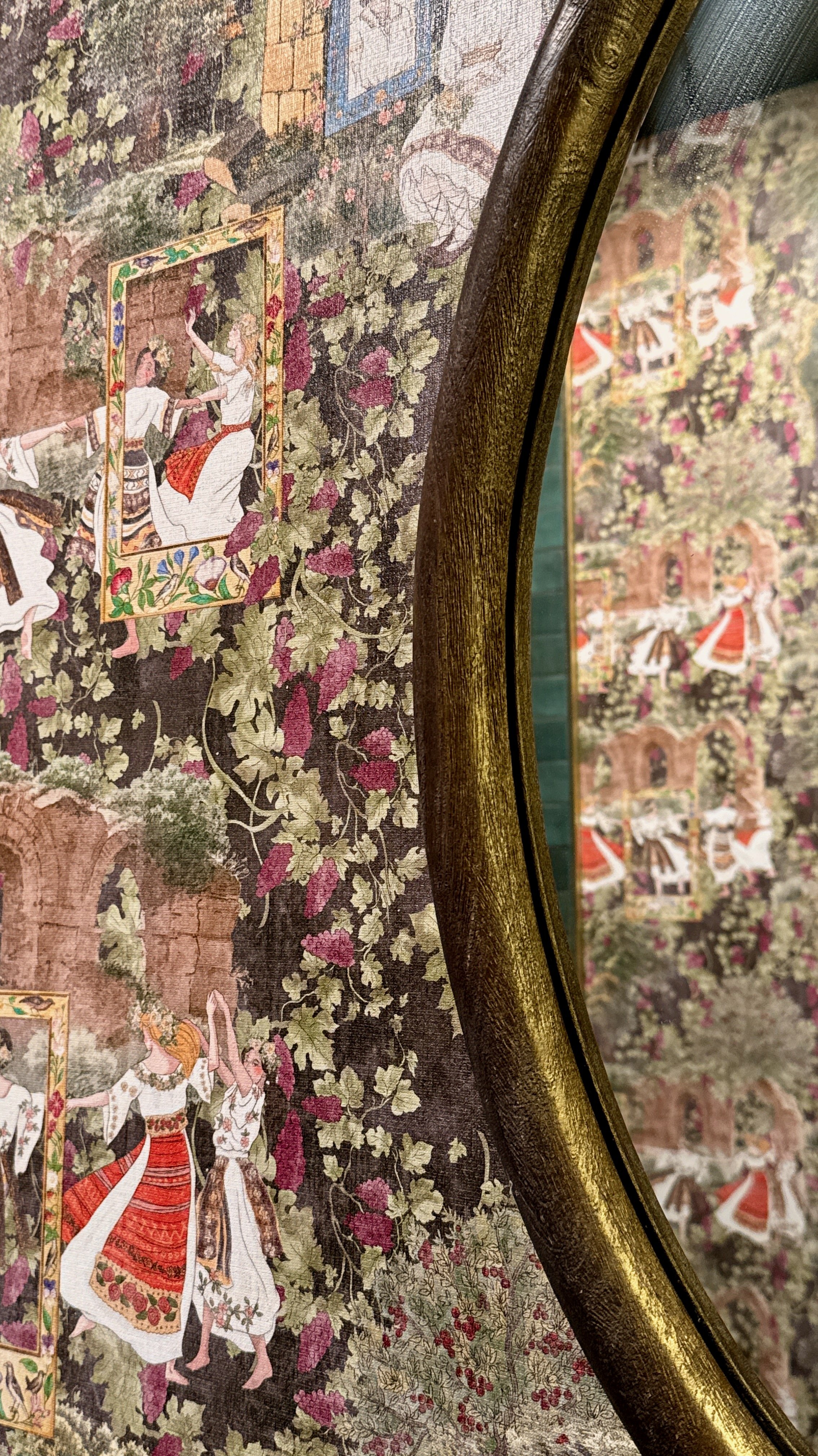 Close-up of a decorative textile with a scene of people in traditional clothing surrounded by floral patterns, seen through a reflective surface with a gold-colored frame.