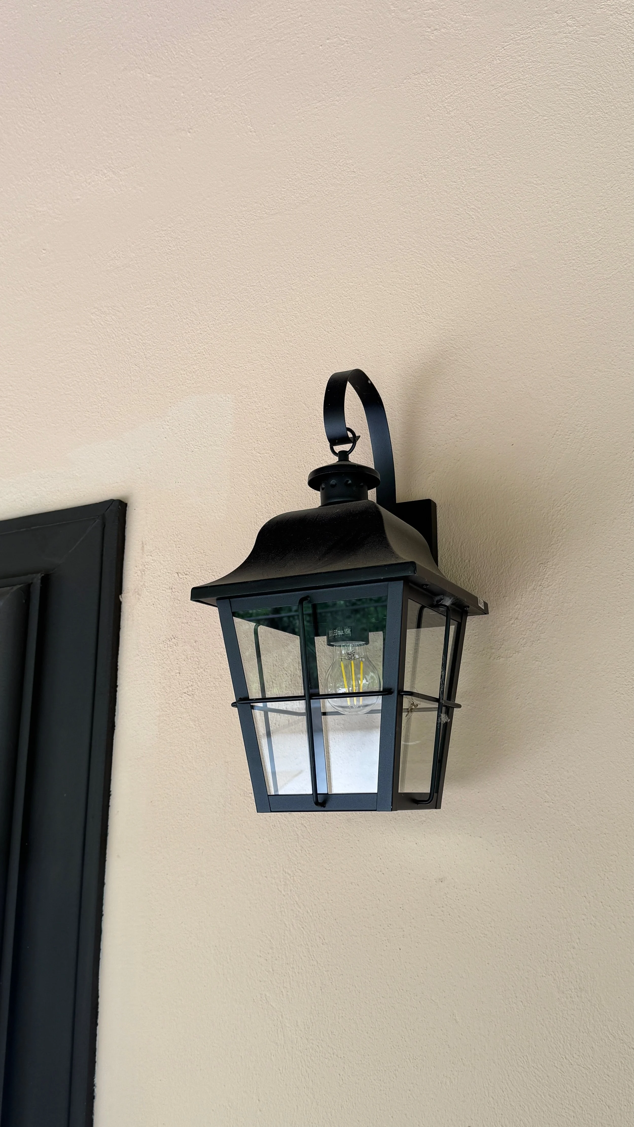 Black outdoor wall-mounted lantern with a glass enclosure and an Edison-style light bulb, mounted on a beige stucco wall.
