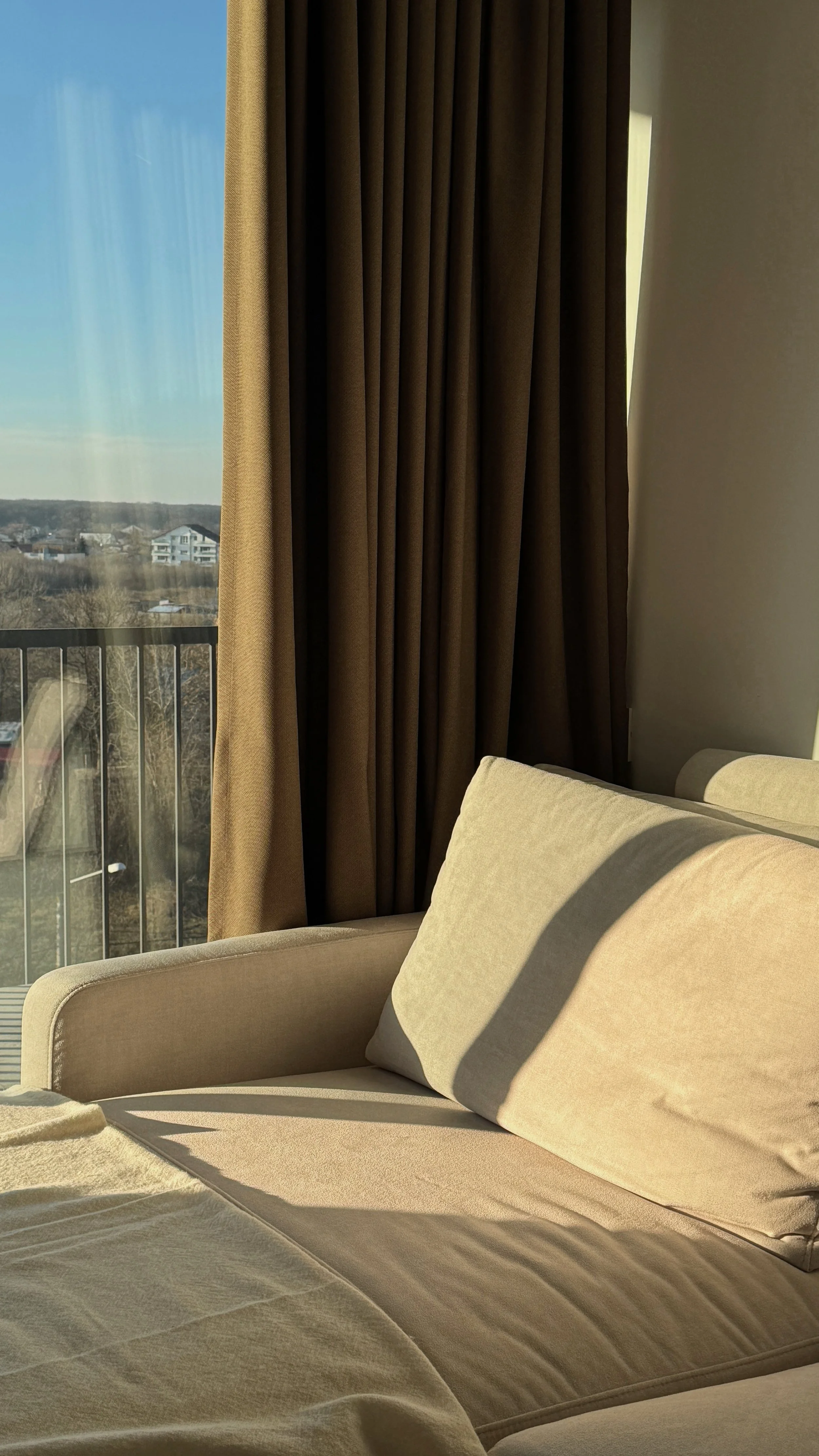 Sunlit beige sofa with cushions next to a window with brown curtains, overlooking a landscape of houses and trees.
