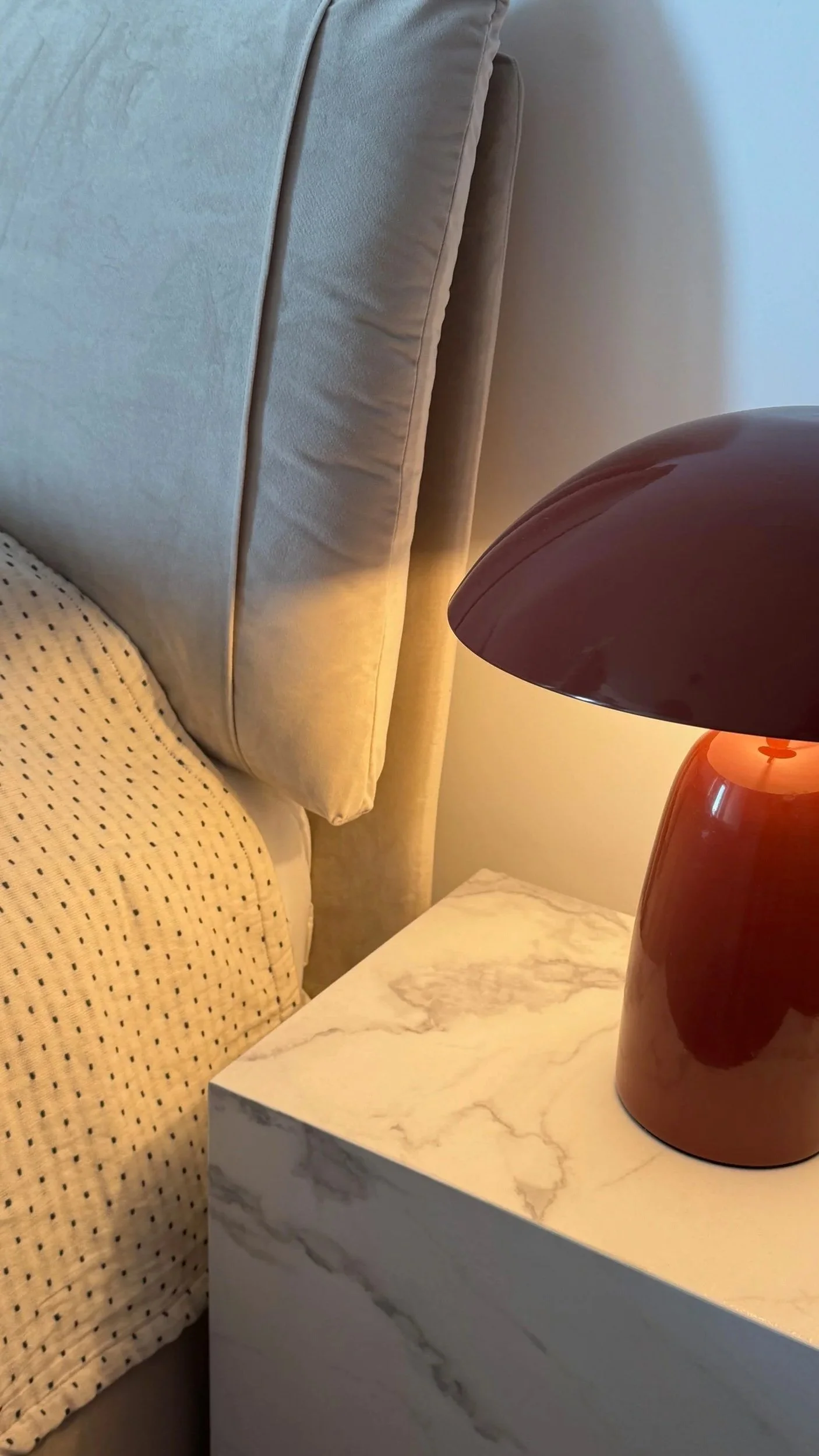 A close-up of a beige upholstered headboard with a beige and black polka dot blanket, a small white marble-top side table with gray veins, and a rust-colored table lamp with a round base and a wide, curved shade.