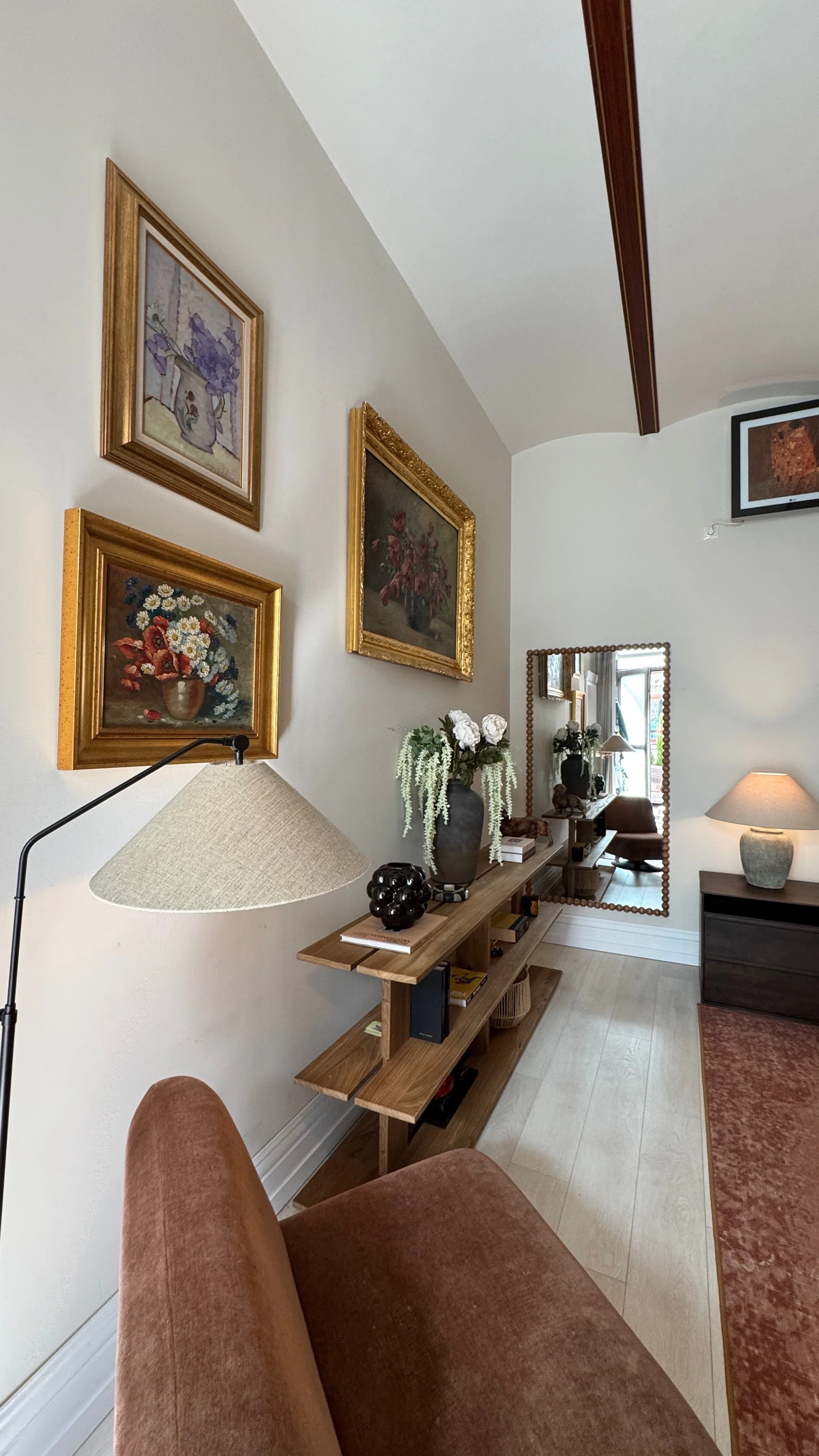 A cozy living room corner with framed floral paintings on the wall, a wooden console table with decorative items and a mirror, and a brown upholstered sofa in front.
