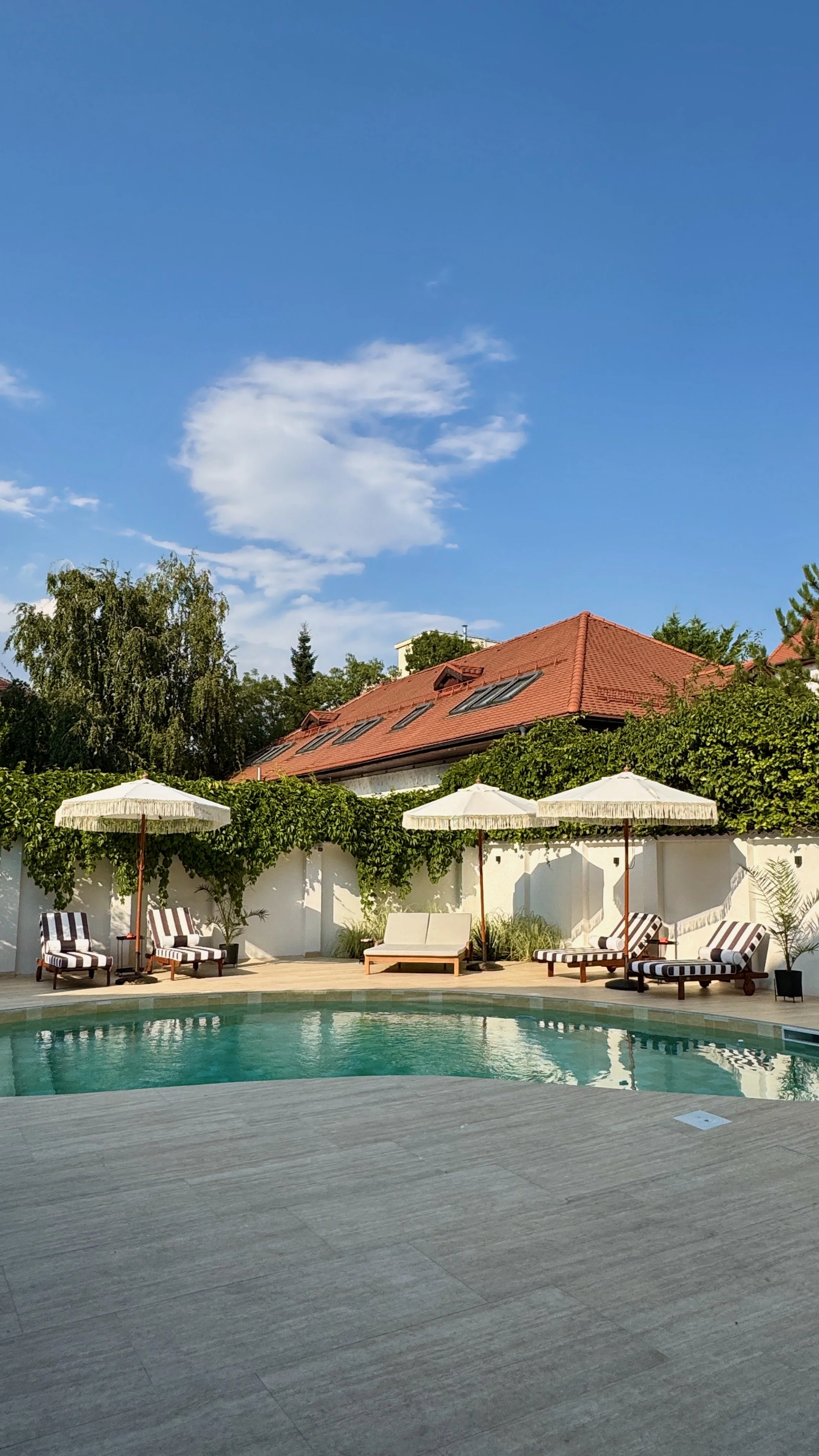 Swimming pool area with lounge chairs, umbrellas, and a white fence, with a house with a red roof and trees in the background under a blue sky with clouds.