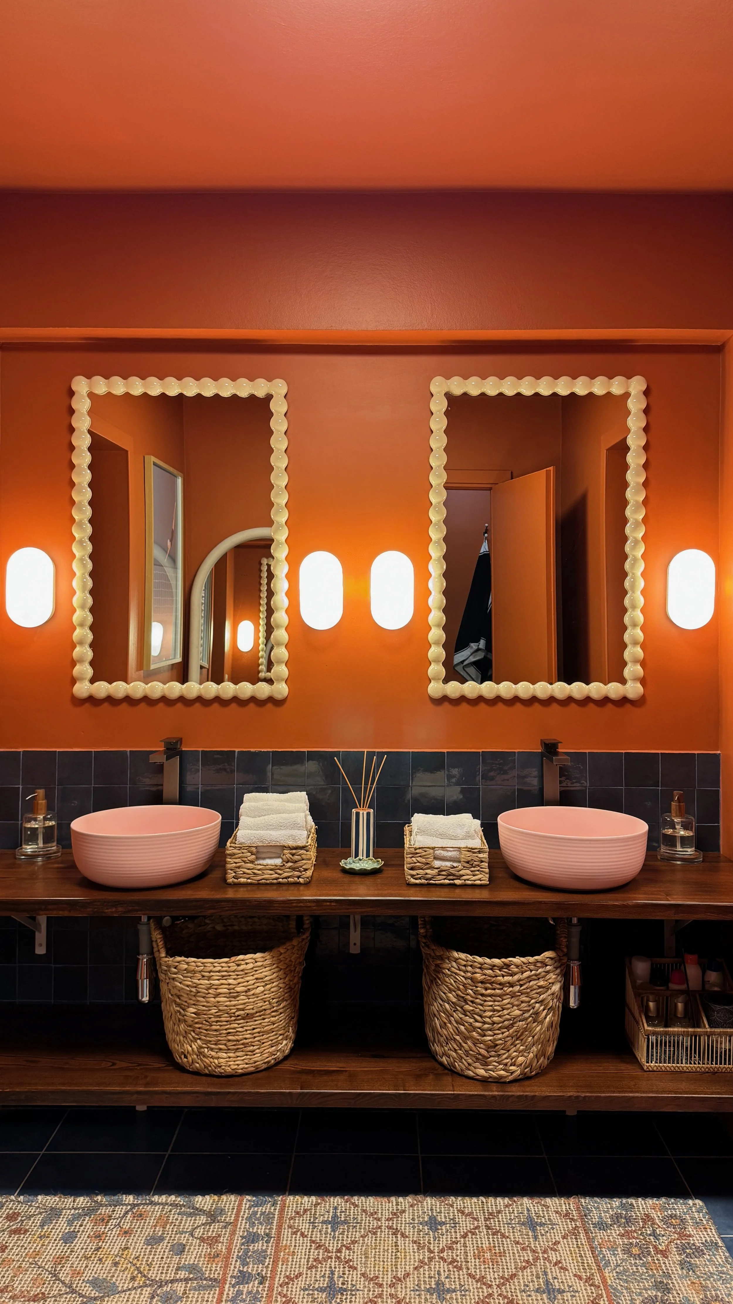 Bathroom vanity with two round pink sinks, black walls, orange wall with two rectangular mirrors with decorative borders, and wicker baskets below holding towels and toiletries, with a patterned rug in front.
