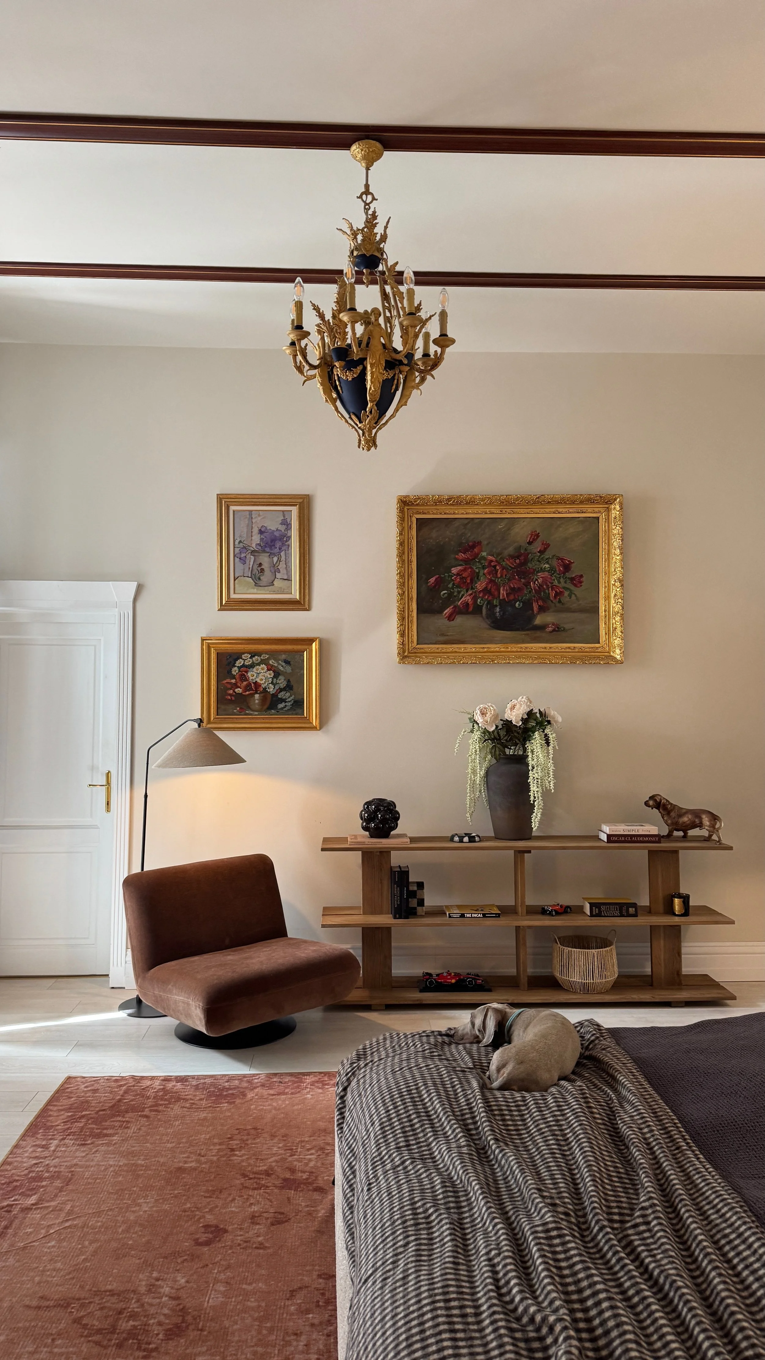 A bedroom with a chandelier, framed artwork, a brown chair, a table with decor, a dog lying on a patterned blanket, and a peach-colored rug.