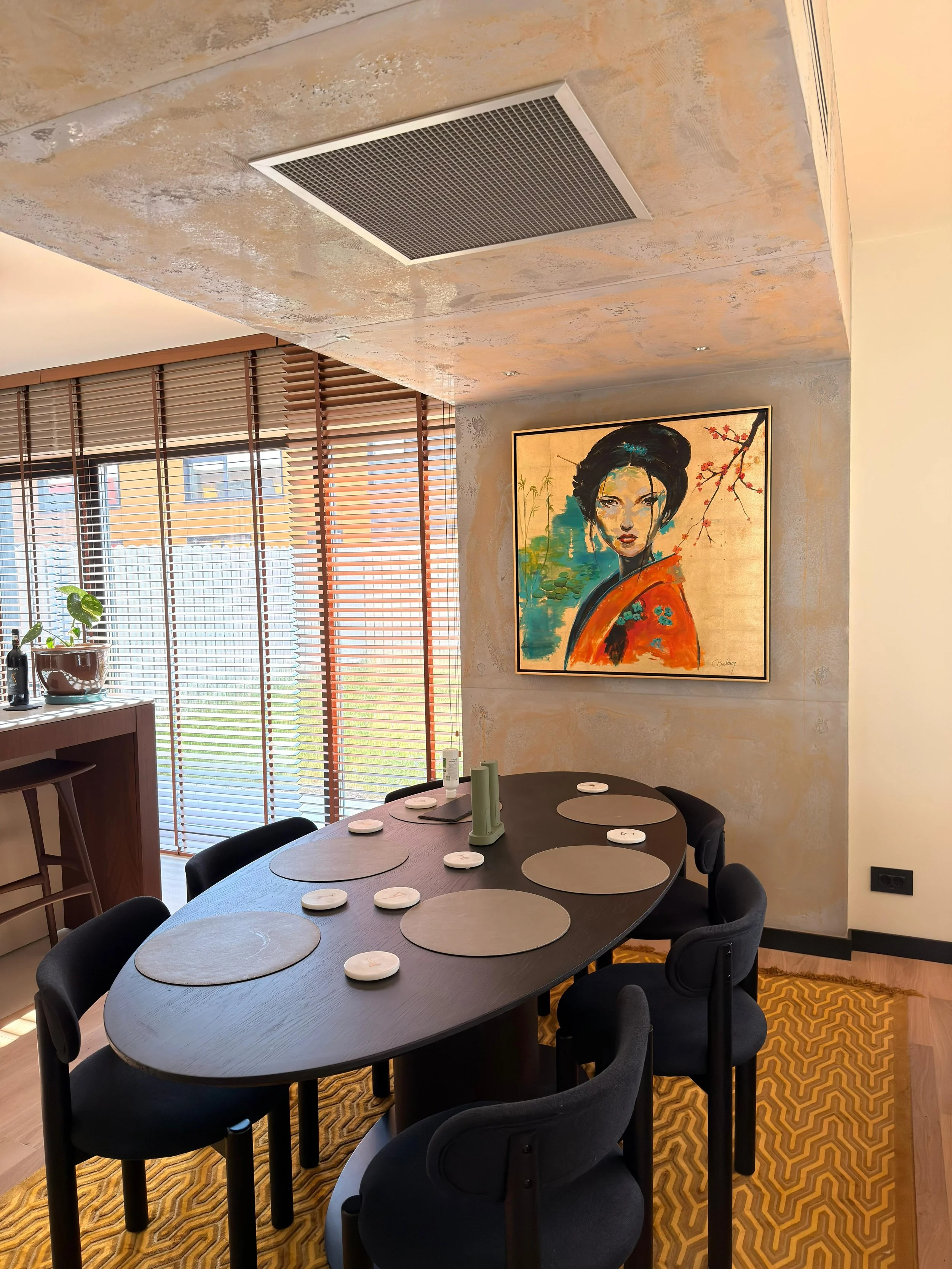A round dining table set for eight, with placemats and small white dishes, surrounded by black chairs in a dining room with a textured wall, a large colorful portrait of a woman in traditional attire on the wall, and large windows with wooden blinds 