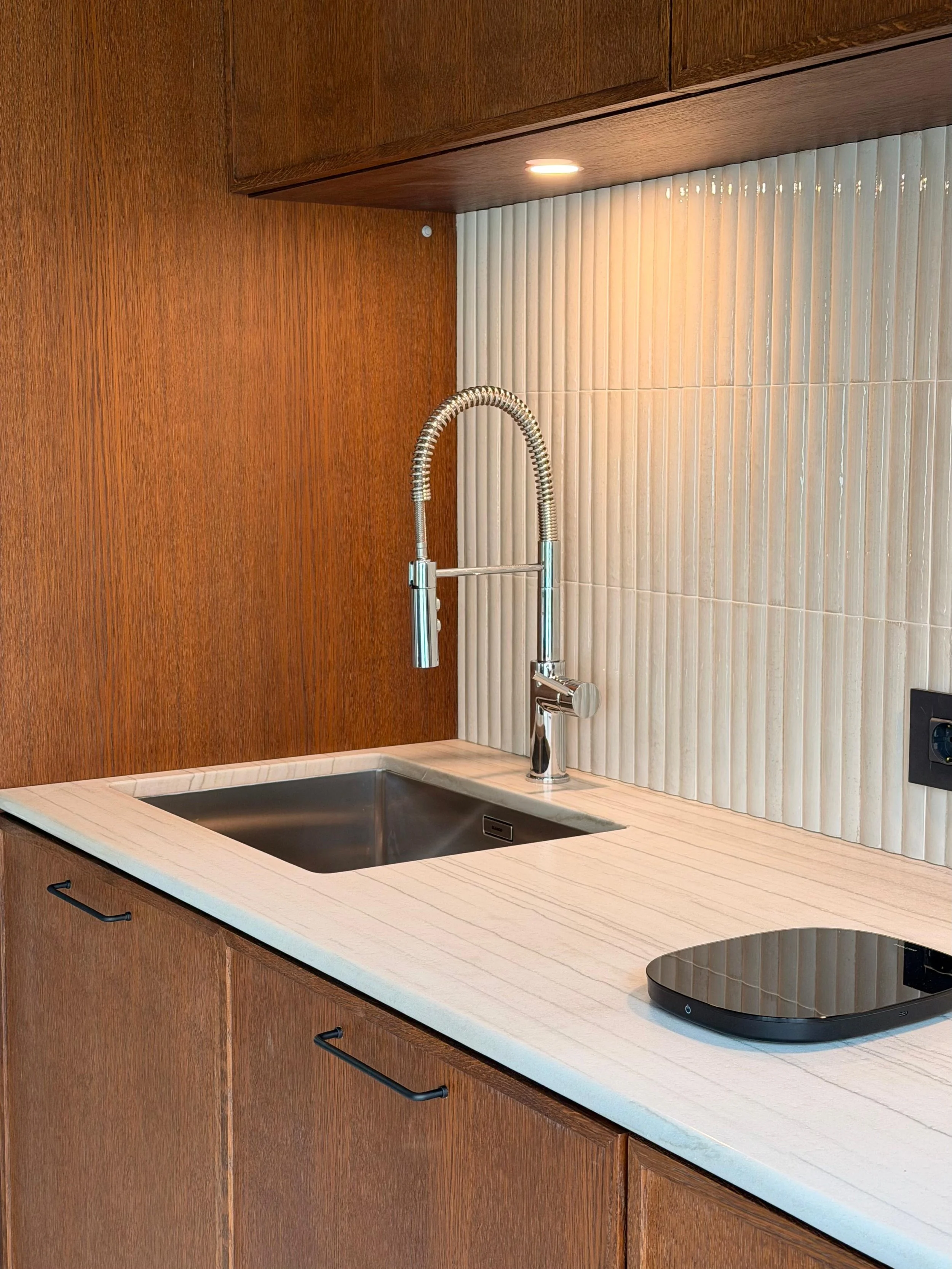 Kitchen counter with a black stovetop, stainless steel sink, and a modern chrome faucet. The background has wood cabinets, a white counter surface, and a beige vertical tile backsplash, with a small recessed light above.