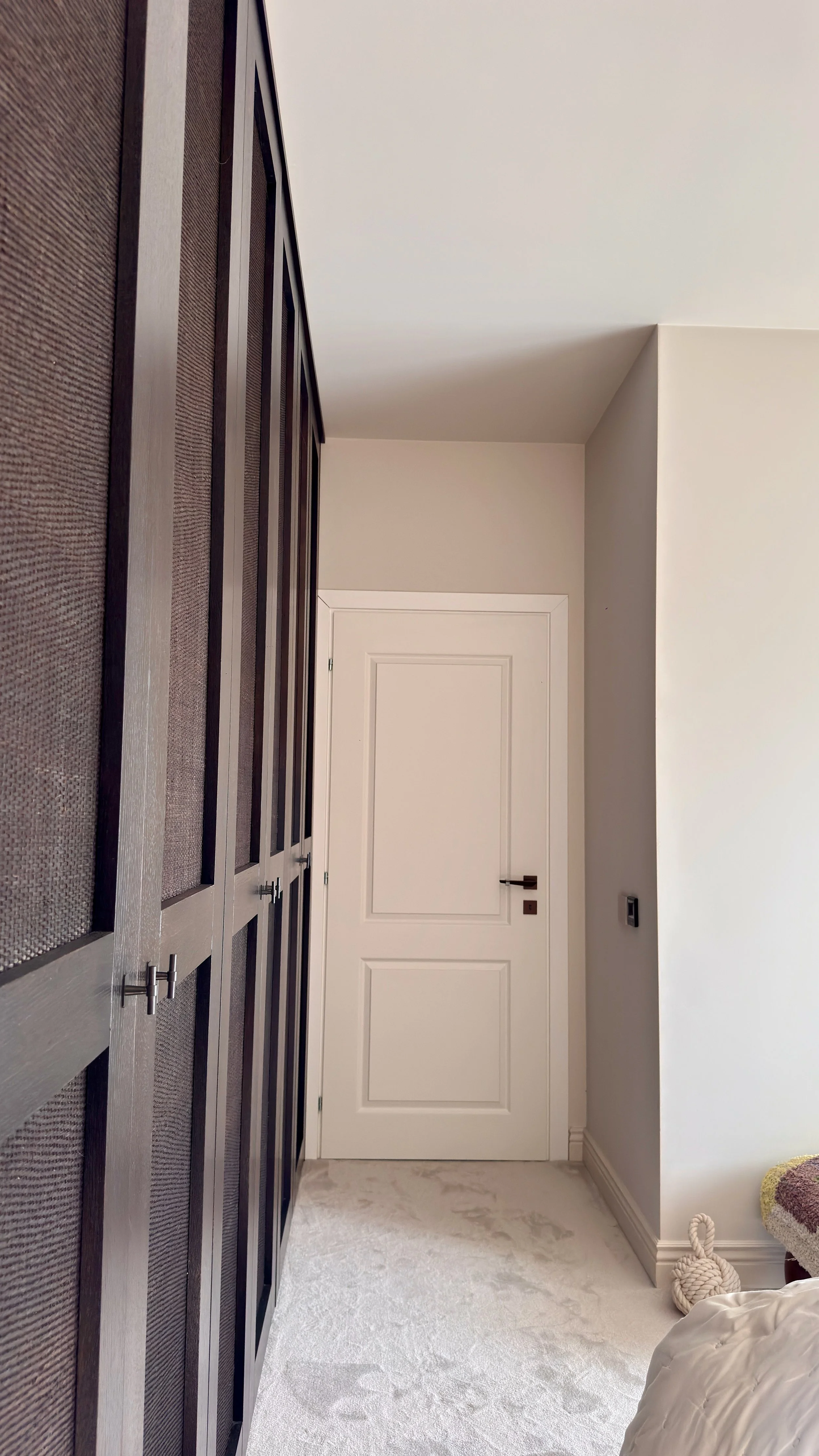 A hallway with a white door, white walls, beige carpet, and a built-in cabinet with glass doors on the left.