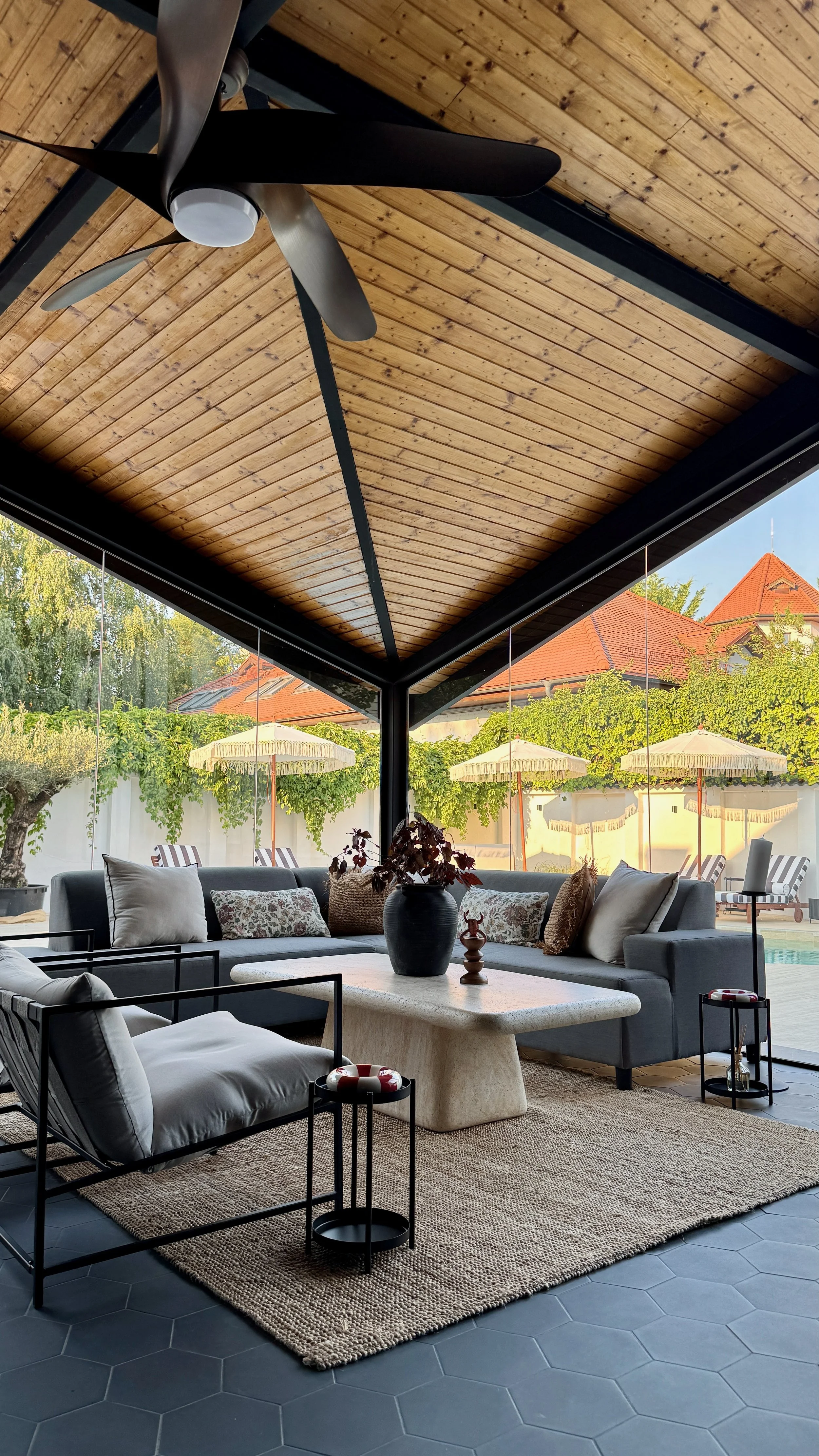 Living room with a large sectional sofa, pillows, a beige rug, a coffee table with a decorative vase, framed by large glass windows showing an outdoor patio with umbrellas and trees, and a wooden ceiling with a ceiling fan.