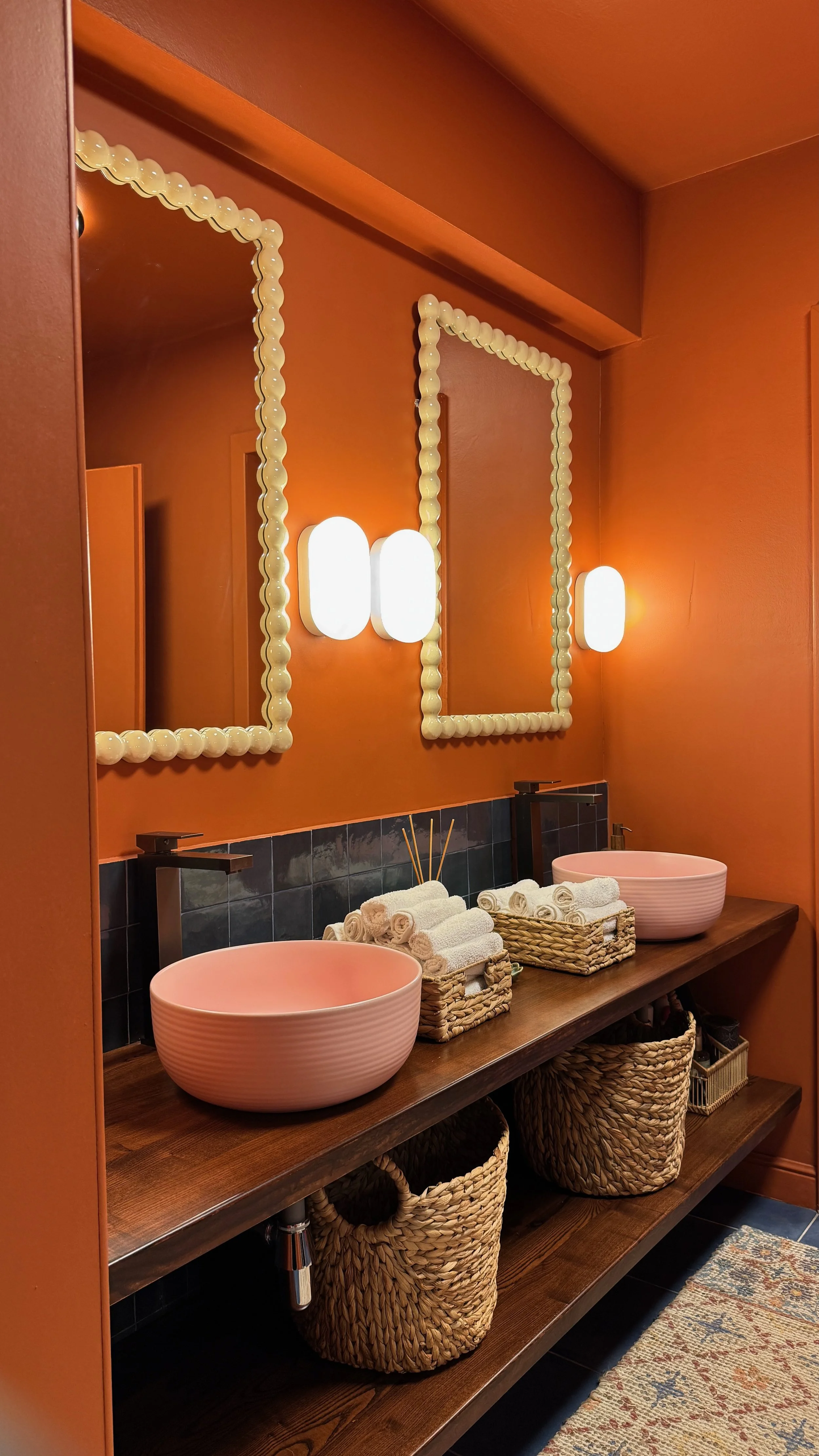 A bathroom vanity with two pink vessel sinks, black faucets, and round towels arranged on the wooden counter. There are two framed mirrors with pearl-like borders, warm orange walls, wall-mounted lights, baskets underneath, and a patterned rug on the