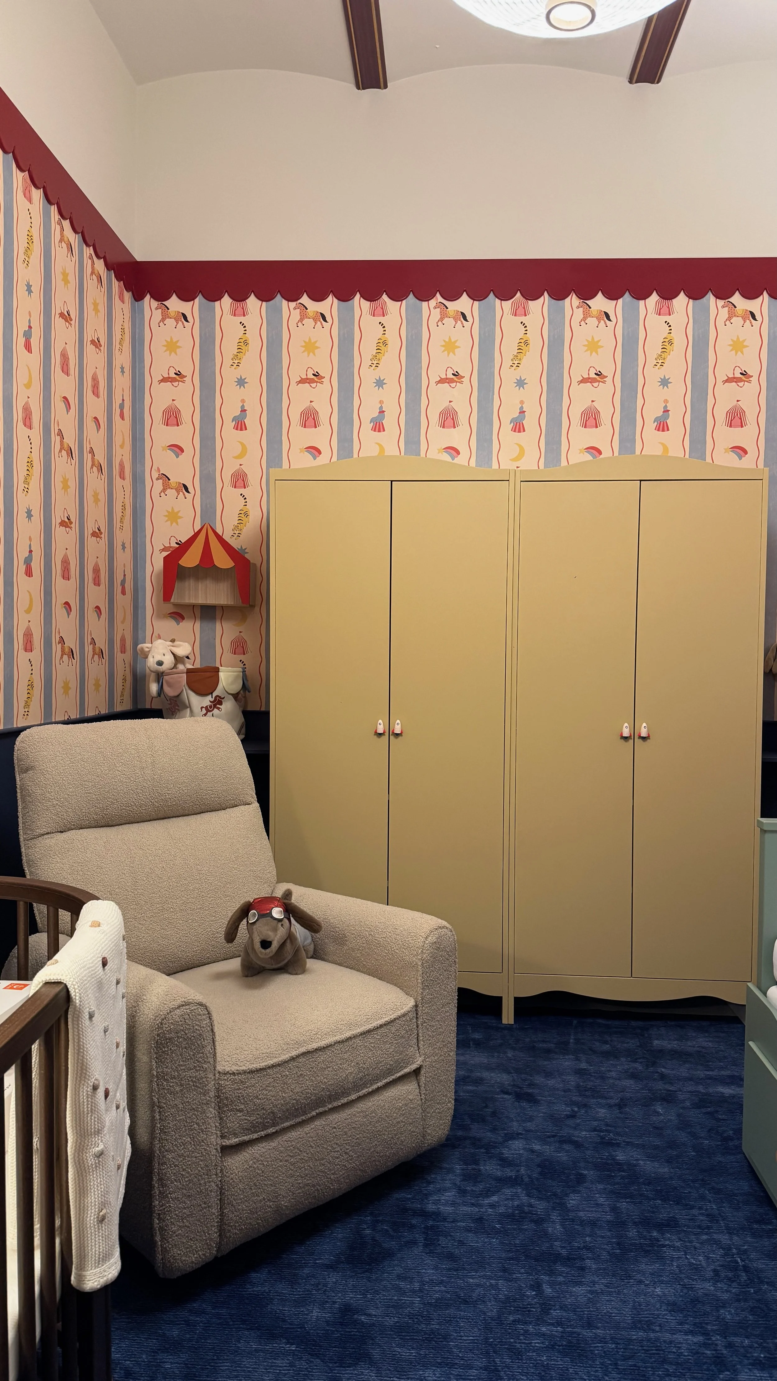 A cozy nursery corner with a beige armchair, a plush dog toy with glasses on the chair, a yellow wardrobe with white handles, a whimsical wallpaper with circus animals and stars, and a corner shelf with a plush cow toy. The ceiling has a white drum-s