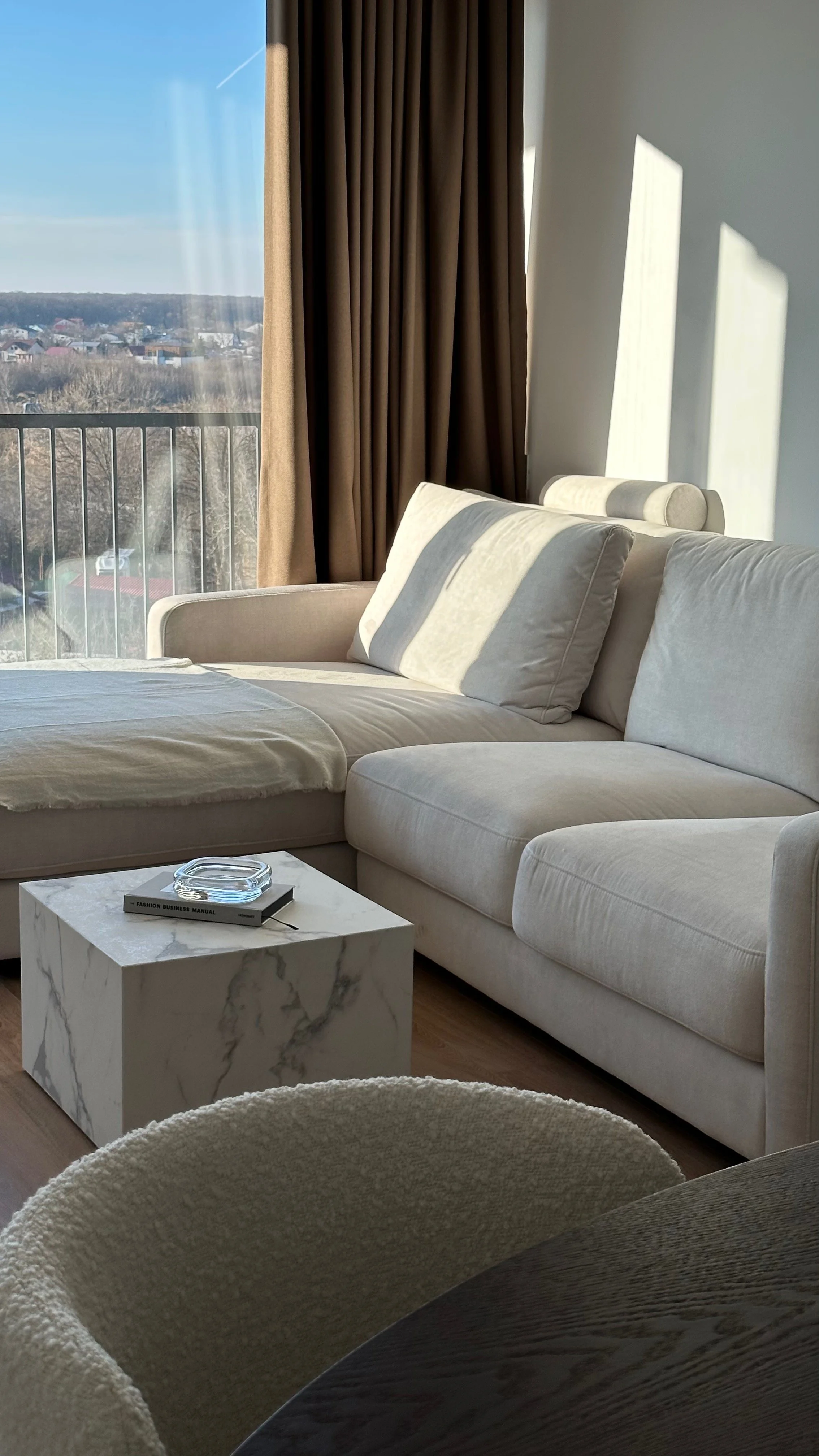 Living room with cream sectional sofa, marble coffee table, and a view of a cityscape outside large window with beige curtains, sunlight casting shadows.