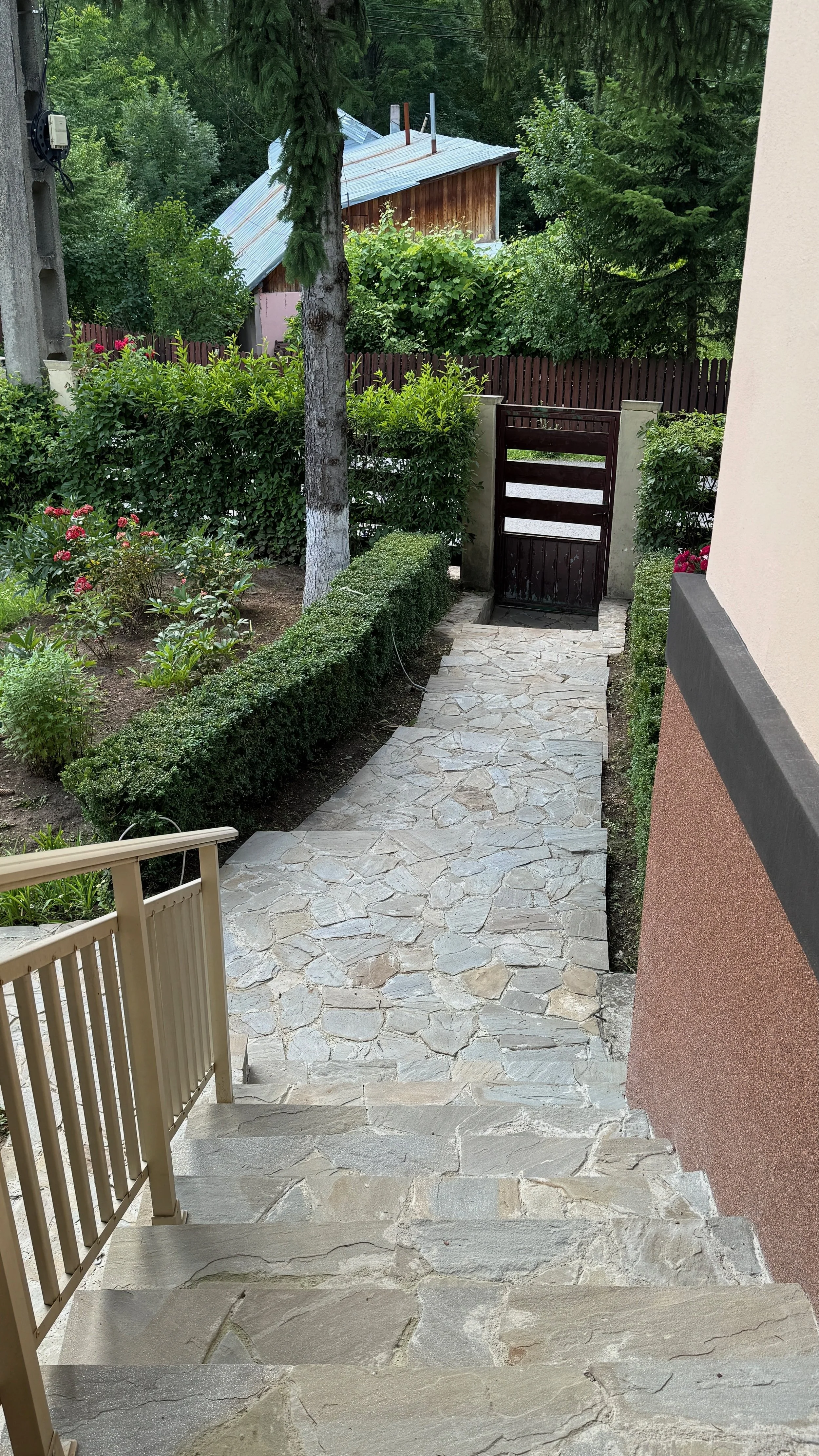 Stone pathway leading to a wooden gate, flanked by greenery and a garden with bushes and flowering plants, with trees and a shed in the background.