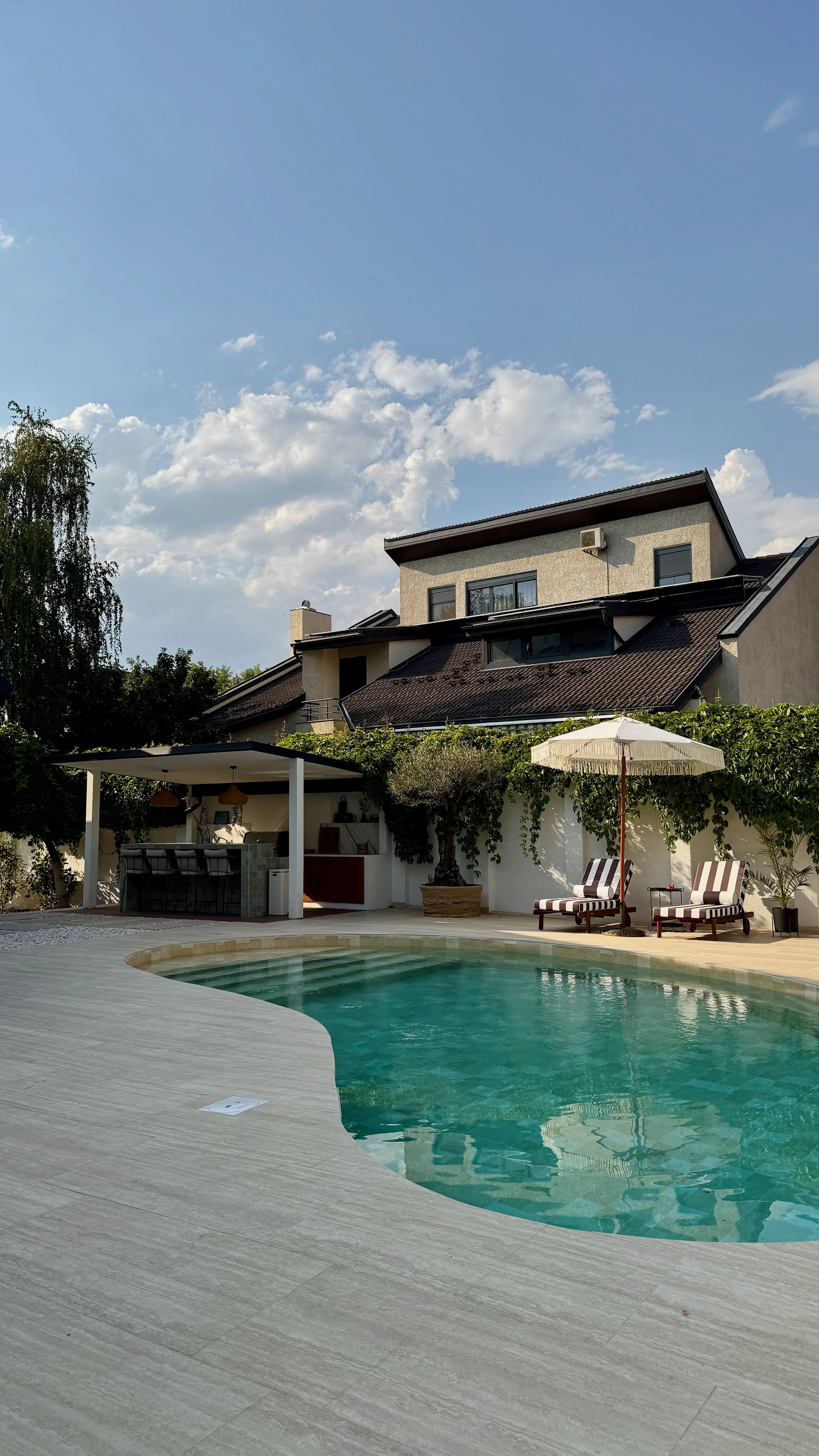 A backyard with a swimming pool, patio chairs, an umbrella, a small bar area, and a house with multiple levels and a balcony under a partly cloudy sky.
