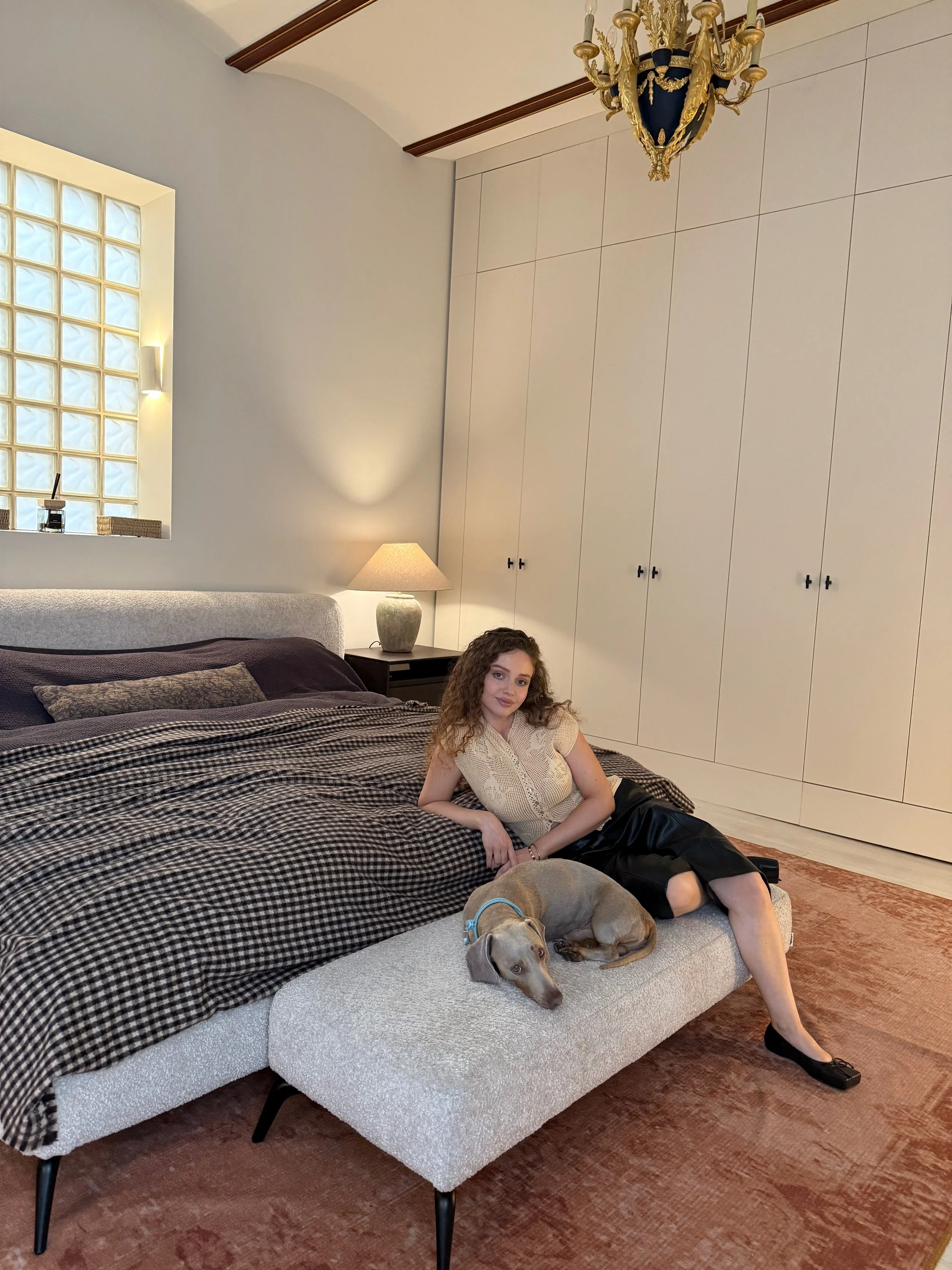 The cuscapes founder sitting on a couch with benny the dog lying next to her. The room has cream-colored walls, a large wardrobe, a lamp, and a chandelier.