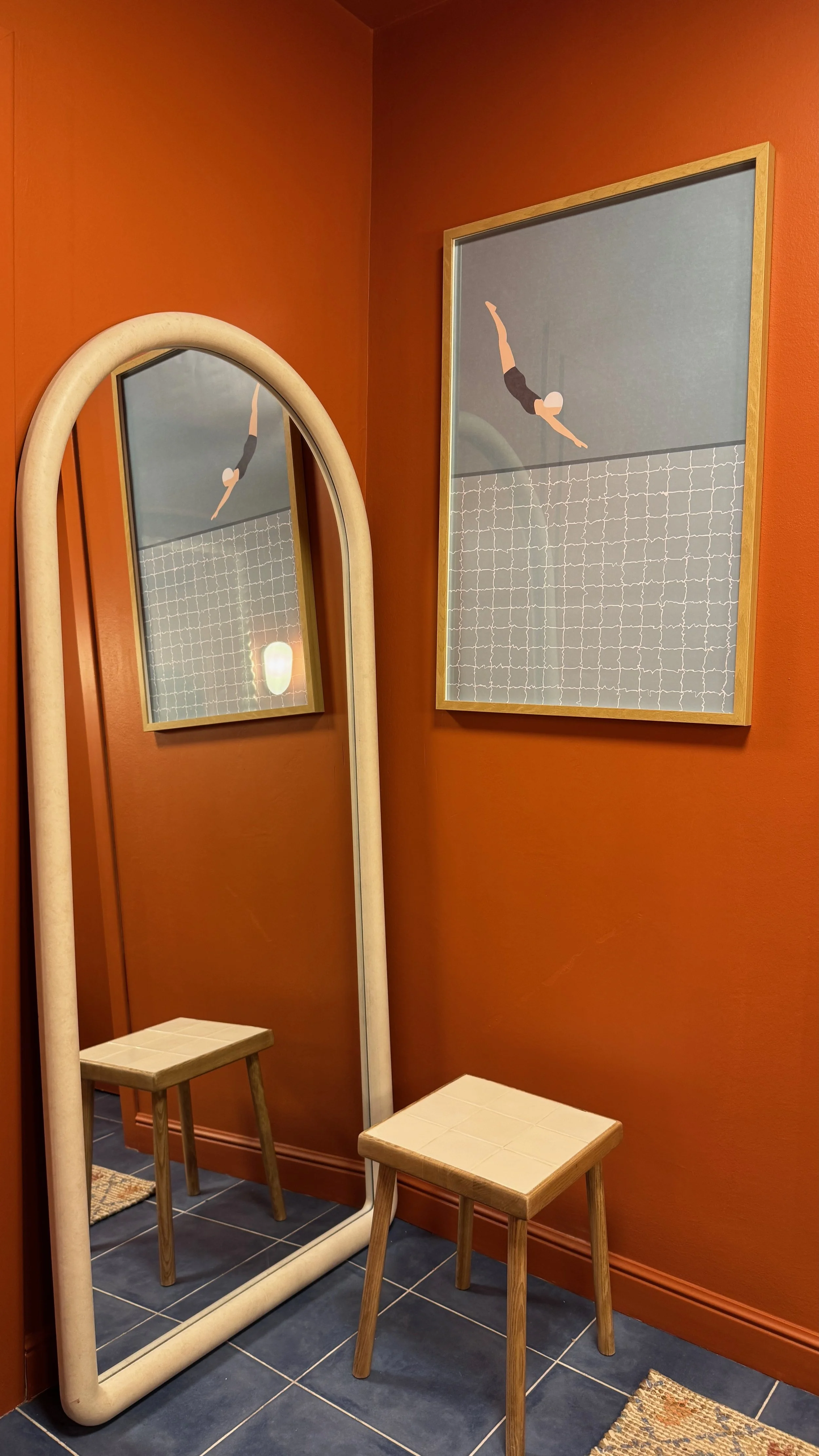 Interior of a room with burnt orange walls, a large mirror with a rounded frame, and a framed artwork of an abstract person diving with a surfboard. Two small wooden stools with tiled tops are in the room. The mirror reflects part of a door, a small 