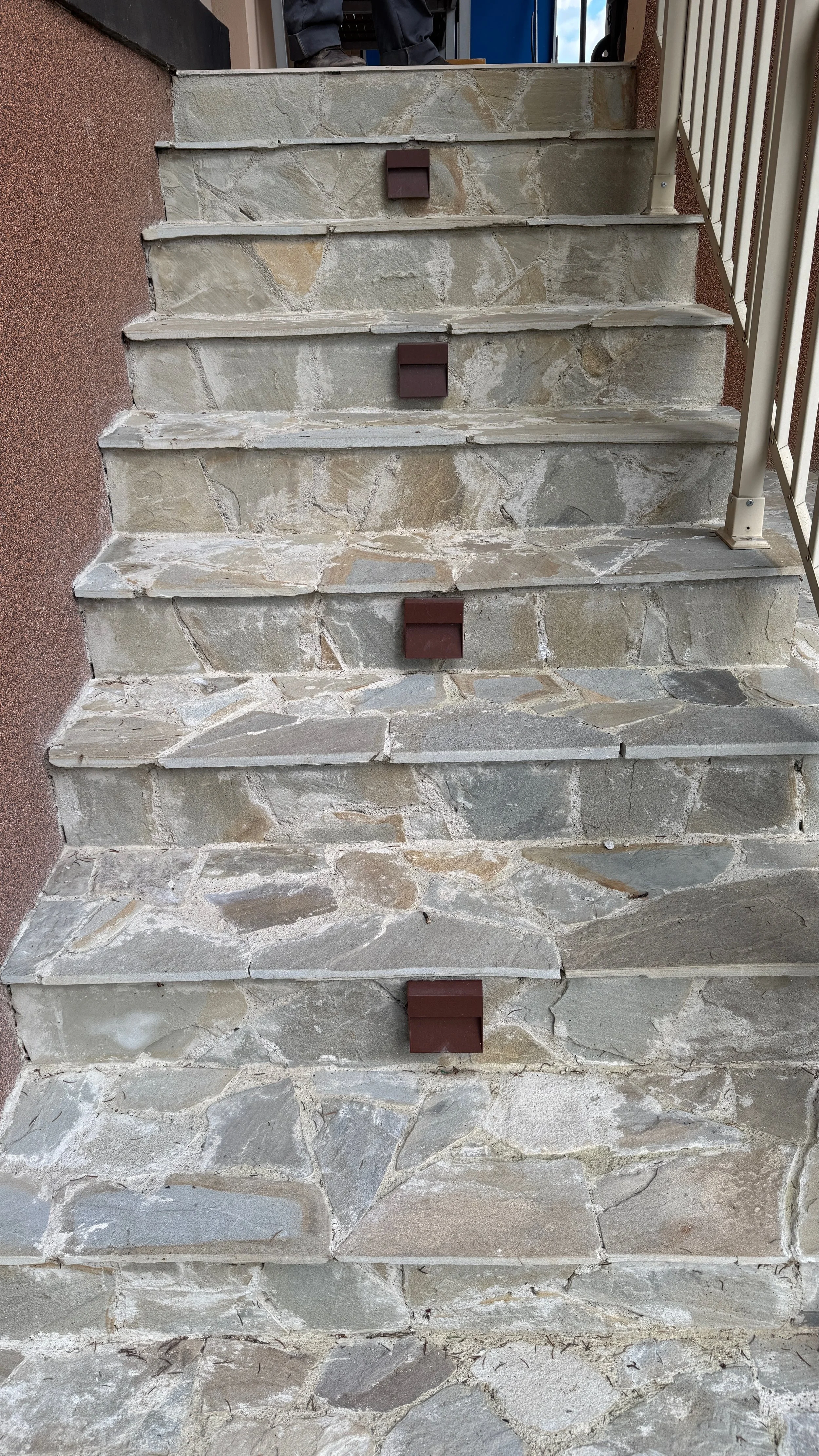 Stone stairs leading up to a doorway or entrance. The stairs are made of large, irregularly shaped stones with mortar in between. There is a metal railing on the right side of the stairs and a brown object at the center of each step.