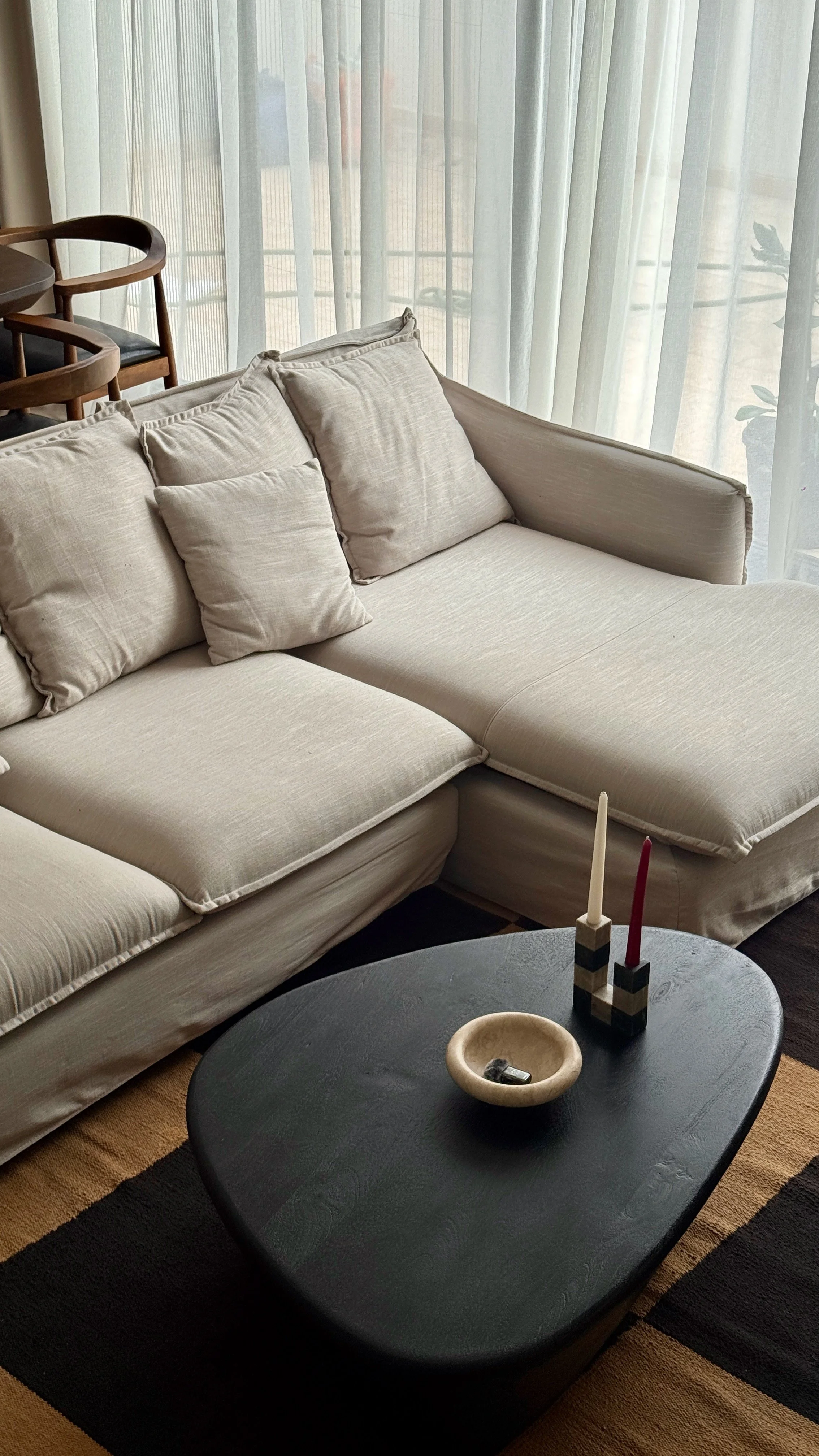 Living room with beige sofa and cushions, a black coffee table with candles and a bowl, and sheer curtains behind, with a dining area with wooden chairs in the background.
