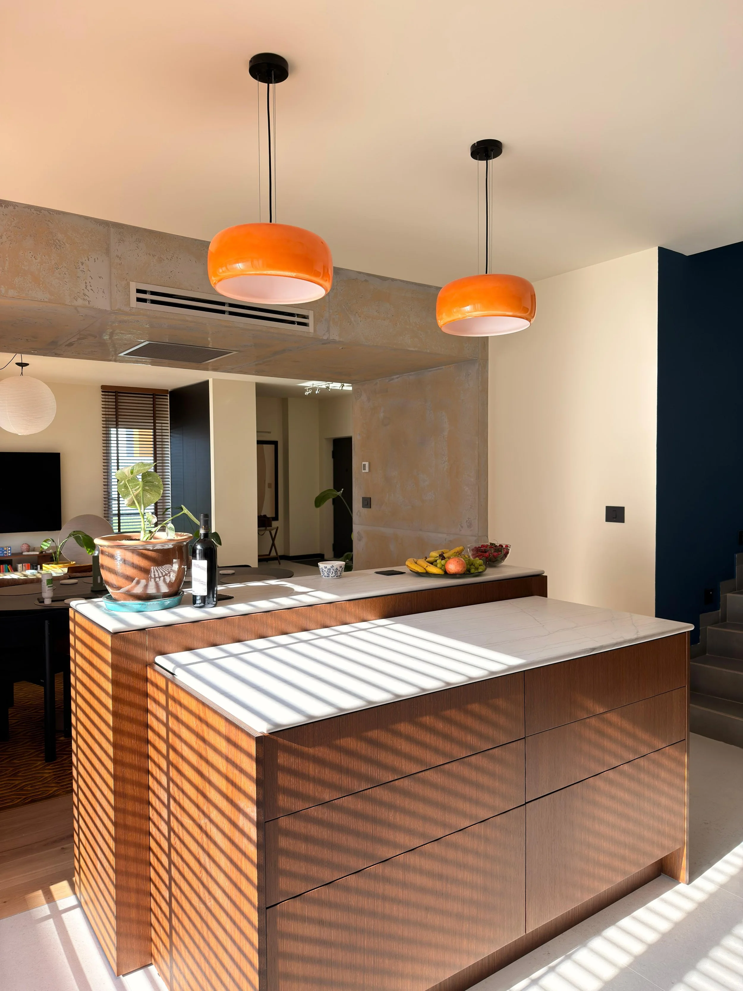 Modern kitchen with a wooden island, white marble countertop, pendant orange lights, indoor plants, and natural light with shadows.