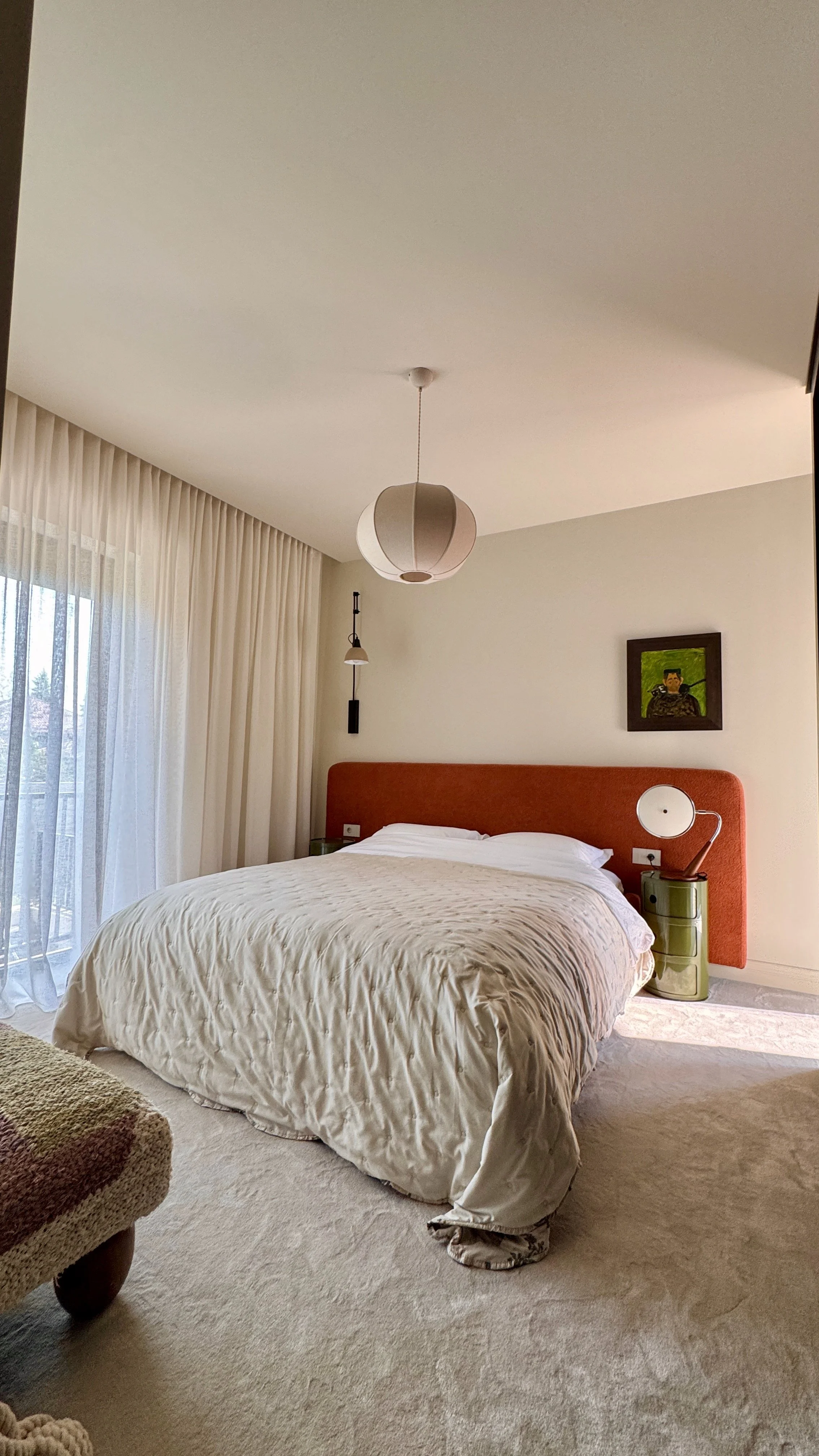 A cozy bedroom with a large bed, white bedding, an orange headboard, a small green side table, and modern lighting fixtures. There are sheer curtains on the window and a framed artwork on the wall.
