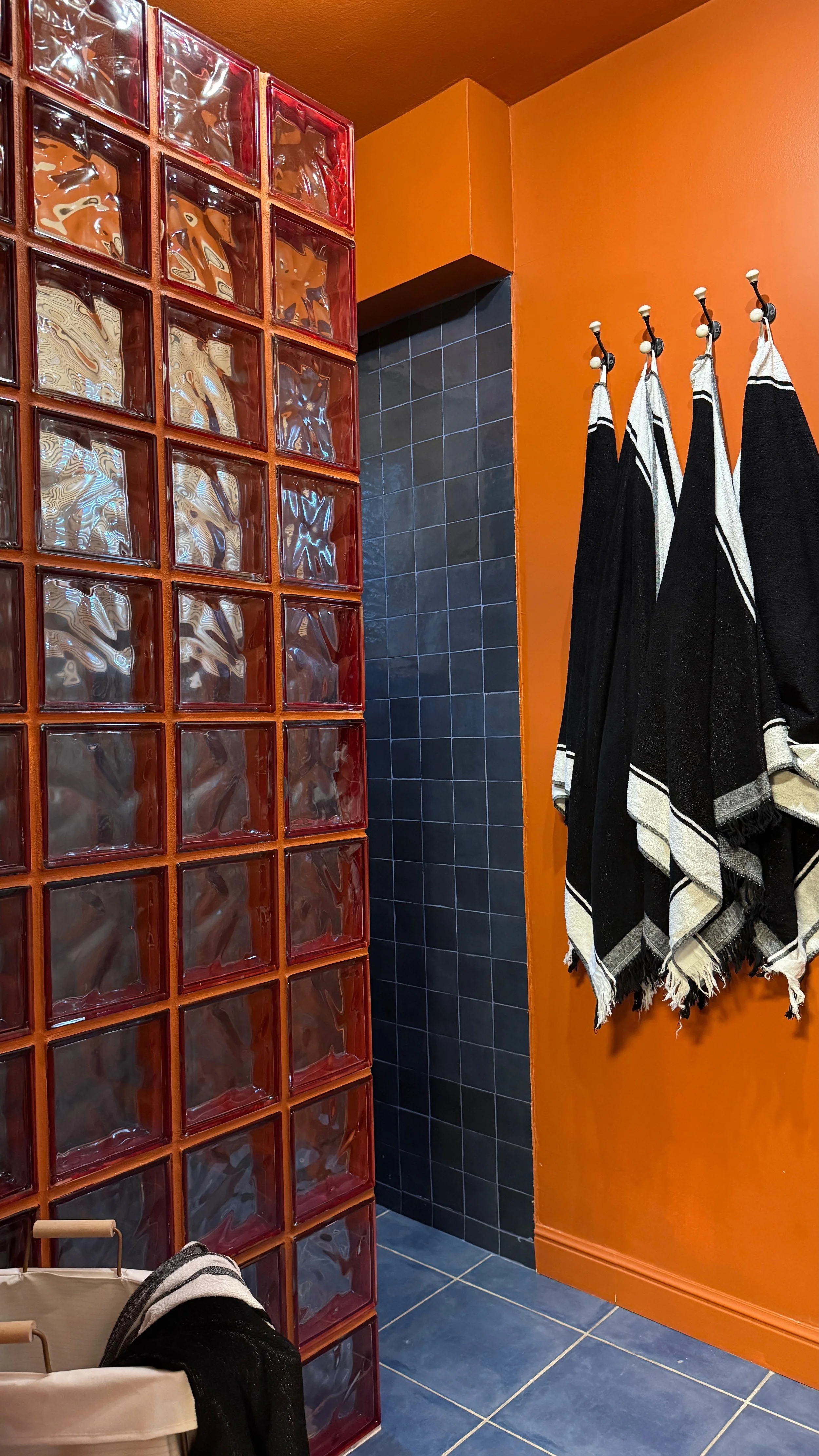 A corner of a room with orange walls and black tiled section. On the right, four black and white towels hang on hooks. On the left, a wall made of glass blocks with a reflective surface.
