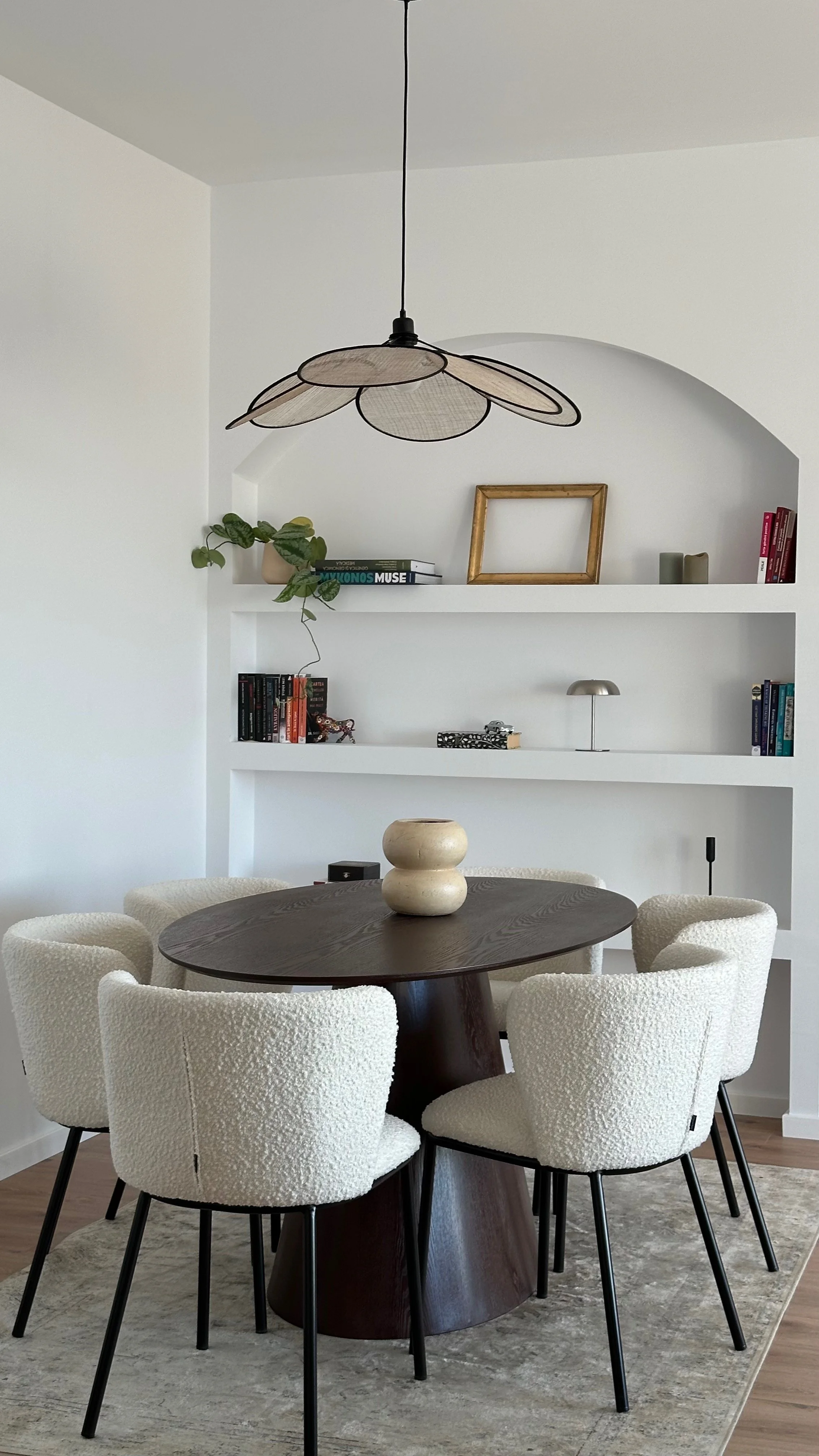 Modern dining room with a round wooden table surrounded by six white upholstered chairs, a decorative vase on the table, a shelving unit with books, a plant, picture frames, and decorative items, and a ceiling light fixture with petal-shaped shades.