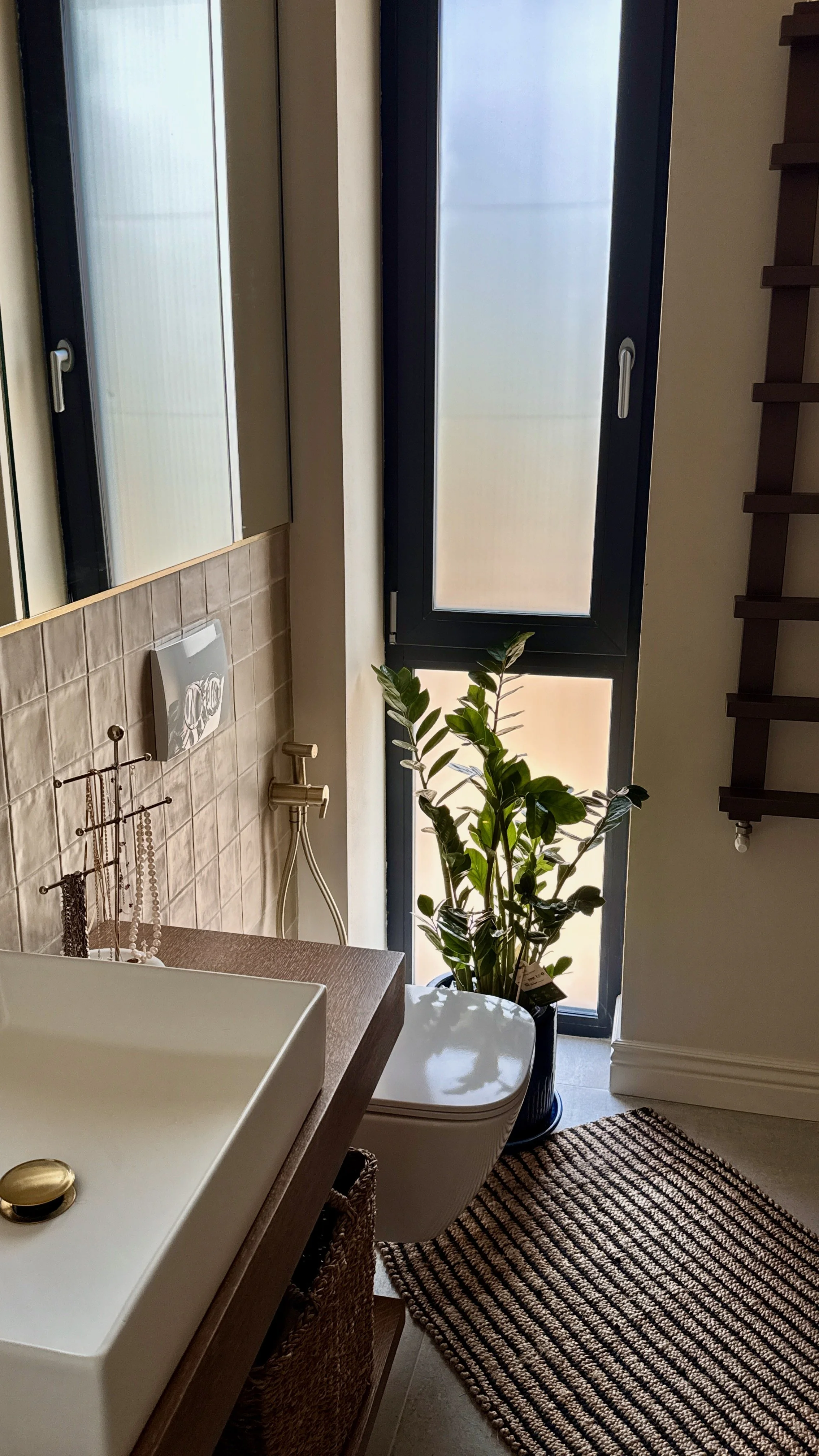 A bathroom with a large window, a potted plant, a countertop with a white vessel sink, a beige tiled wall with a mirror, and a woven rug on the floor.