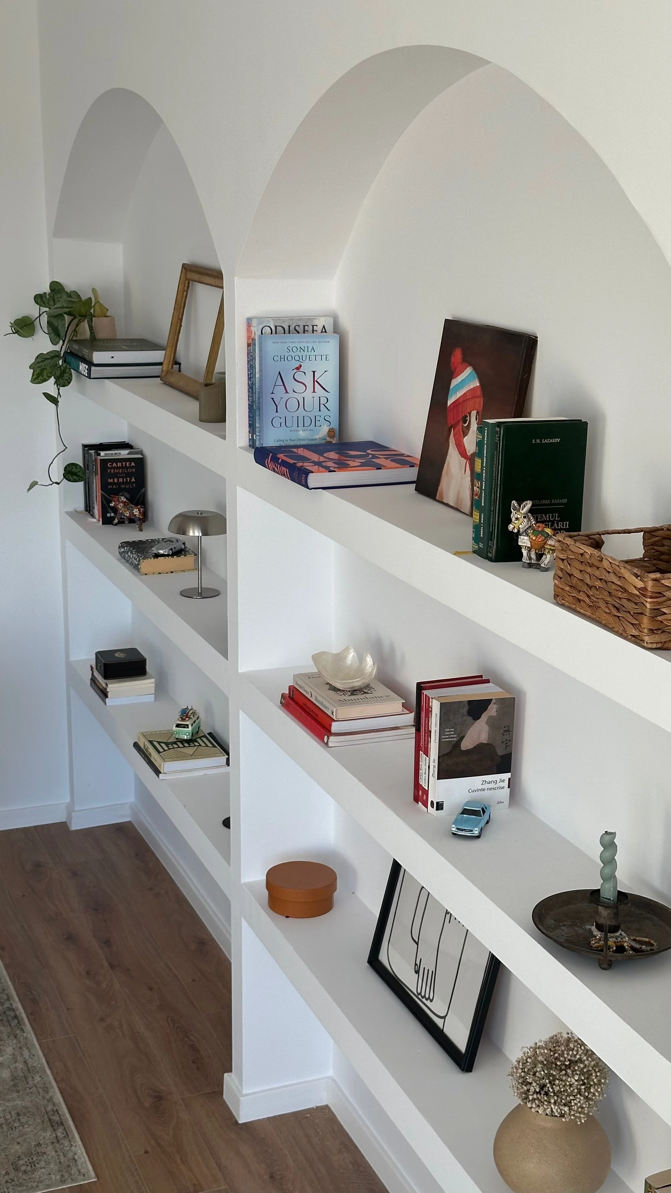 White bookshelf with multiple shelves filled with books, decorative items, and small plants, placed against a white wall with arched spaces.