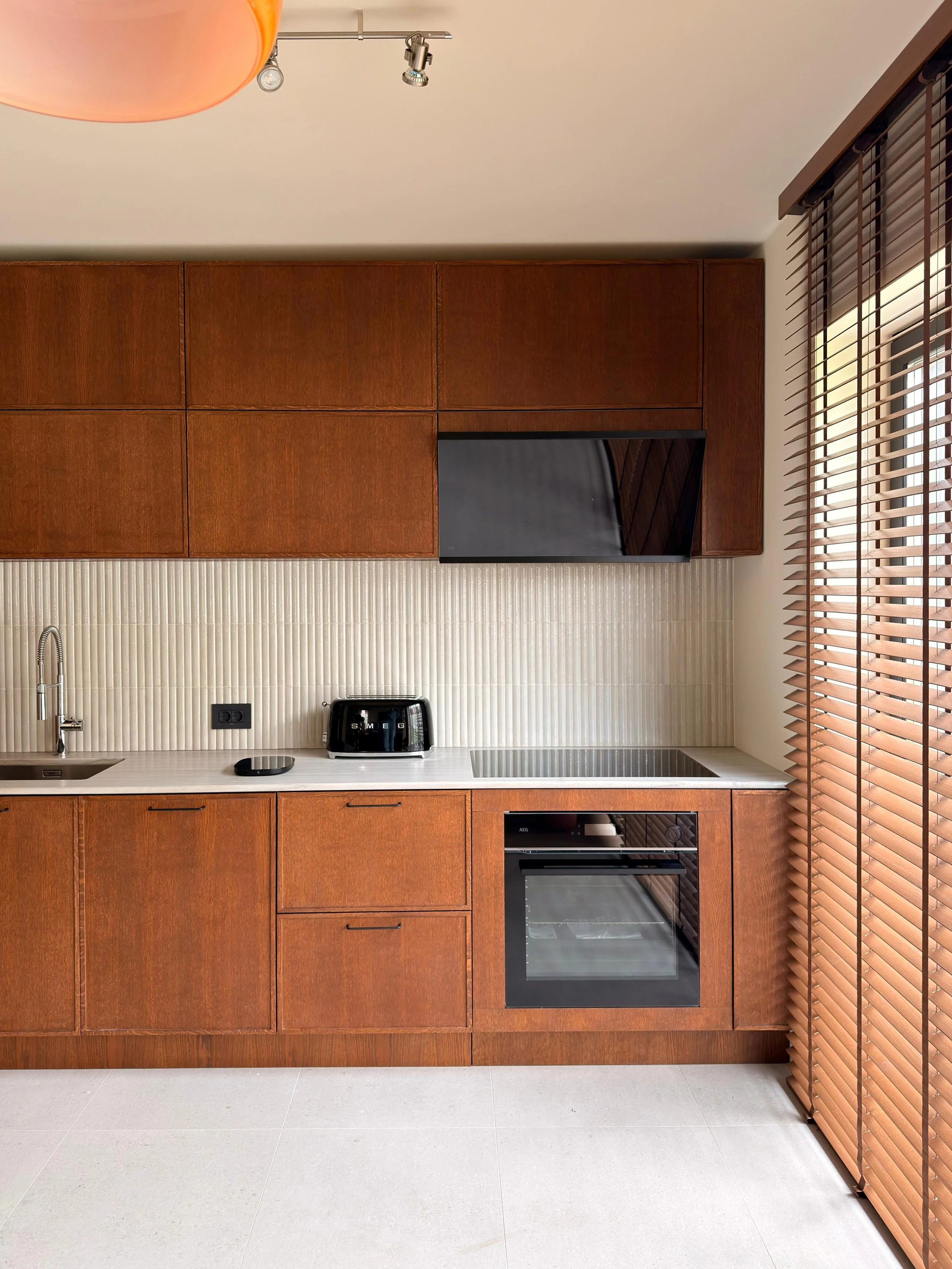 Modern kitchen with wooden cabinets, white countertop, black built-in oven, stovetop, toaster, sink, and window with wooden blinds.
