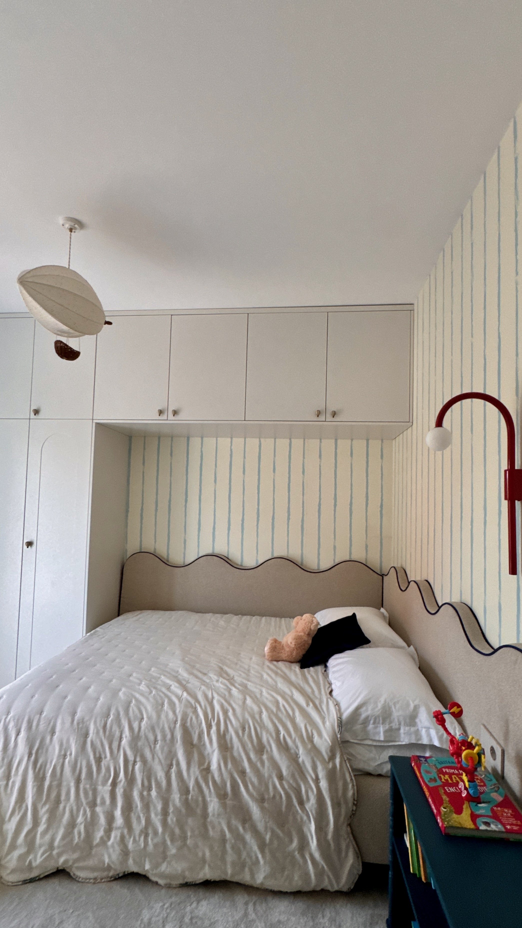 A bedroom with a bed covered by a white quilt, two pillows, and a teddy bear, along with a drawer with children's books and toys. The wall behind the bed has striped wallpaper, and there are built-in white cabinets above the bed. A red lamp is mounte