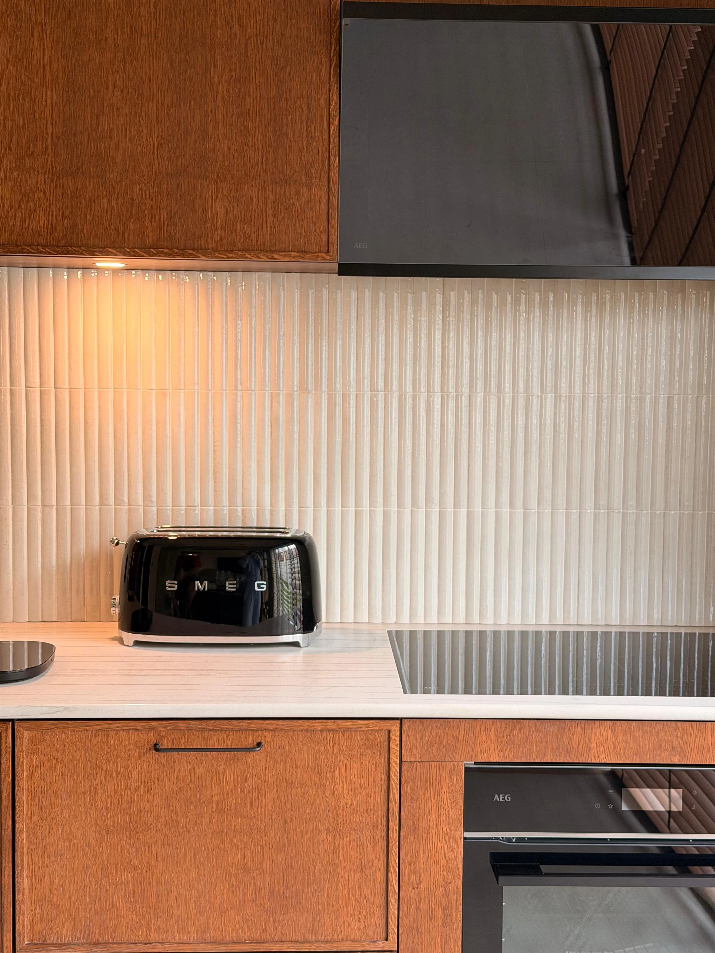 Kitchen countertop with beige tiles, wooden cabinets, a black toaster, and two black AEG appliances, possibly an oven and a microwave.