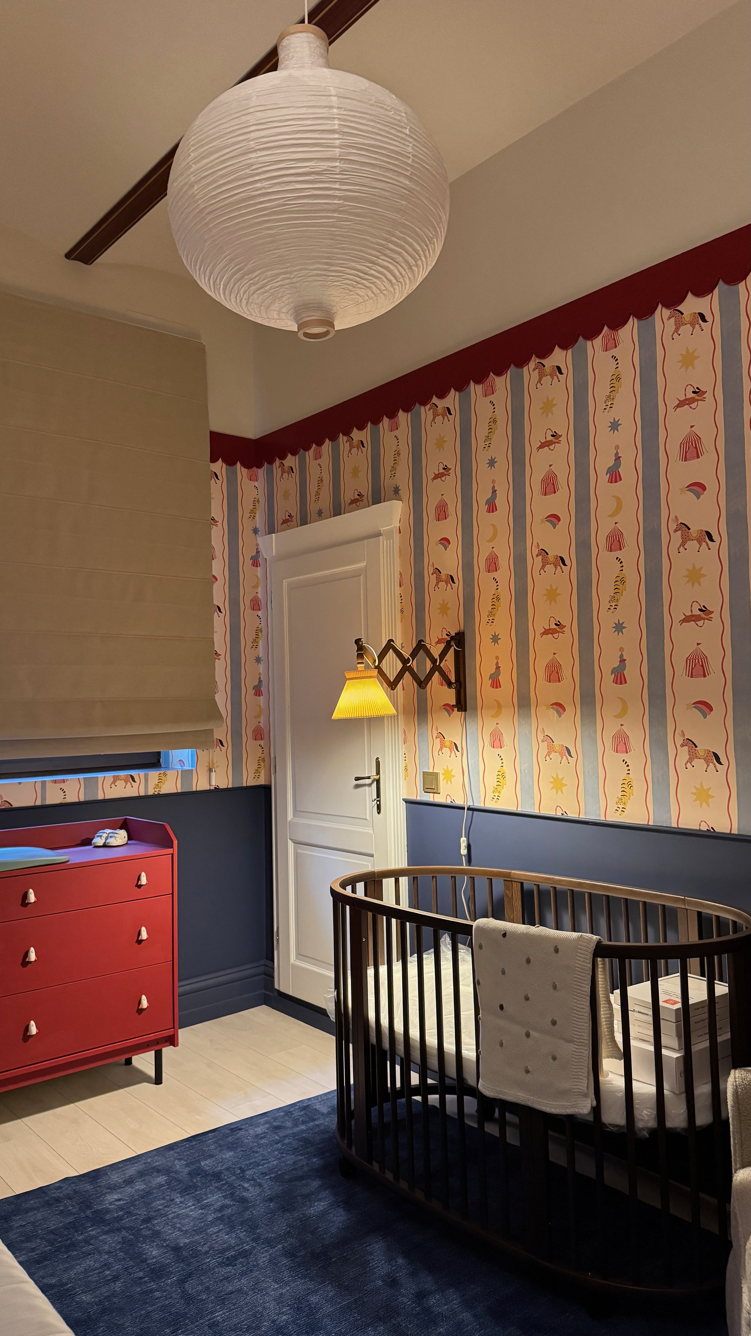 A nursery room with a paper lantern ceiling light, colorful circus-themed wallpaper, a white door with decorative molding, a yellow wall-mounted lamp, a red dresser with white knobs, a black crib with a white mattress, a grey towel hanging on the cri