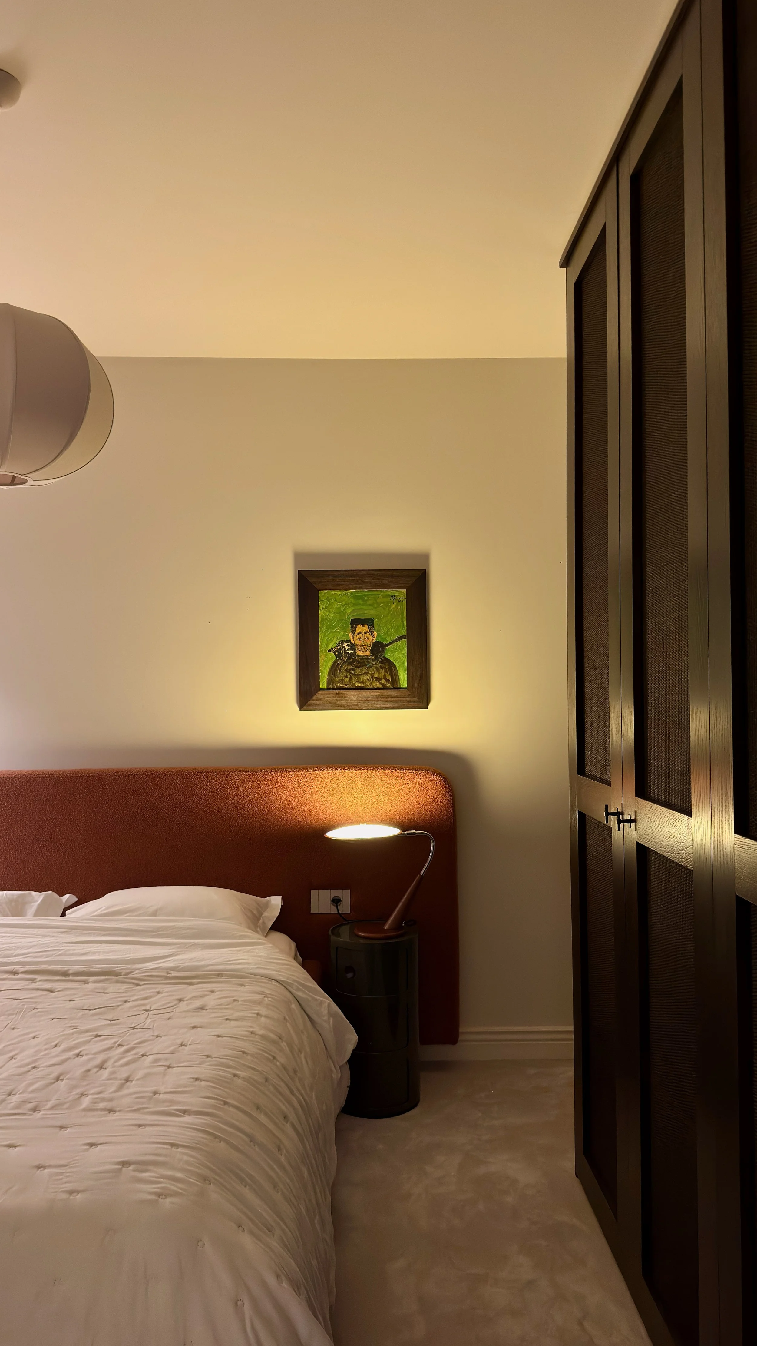 A master bedroom with a bed, a nightstand with a lamp, a framed painting on the wall, and a large wardrobe.