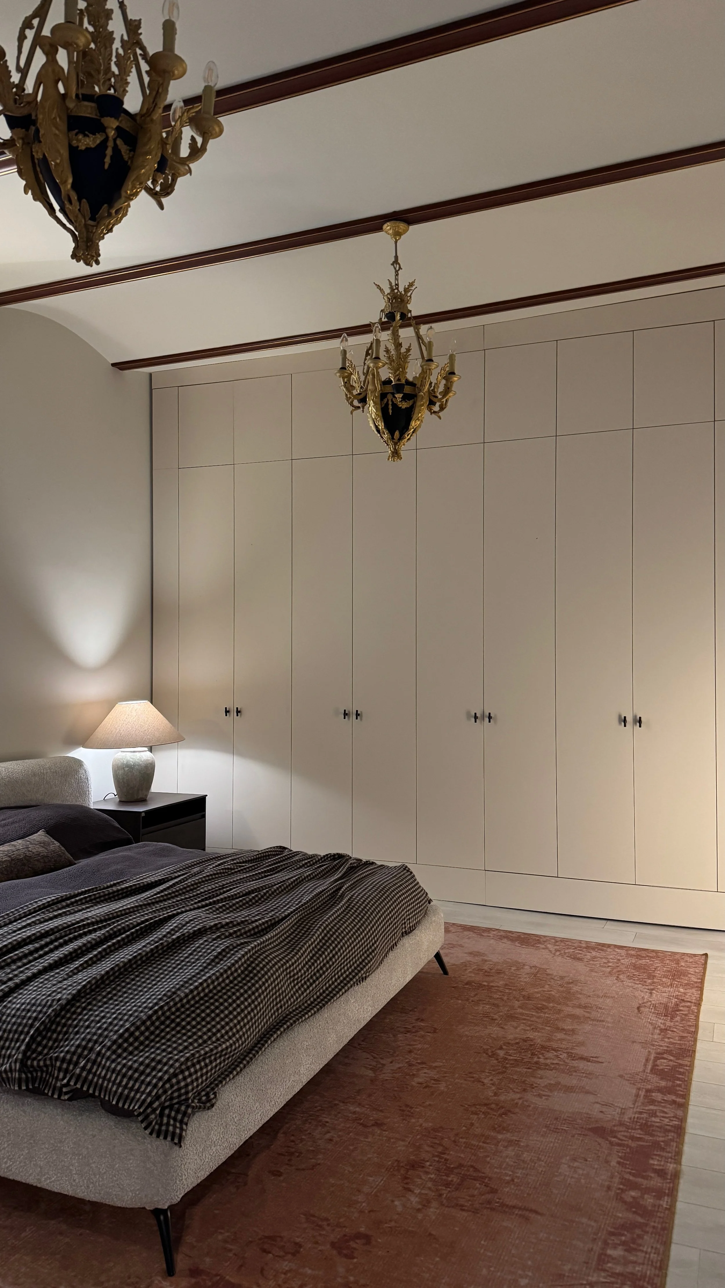 A bedroom with a bed, a lamp on a nightstand, a large white wardrobe, and two chandeliers hanging from the ceiling.