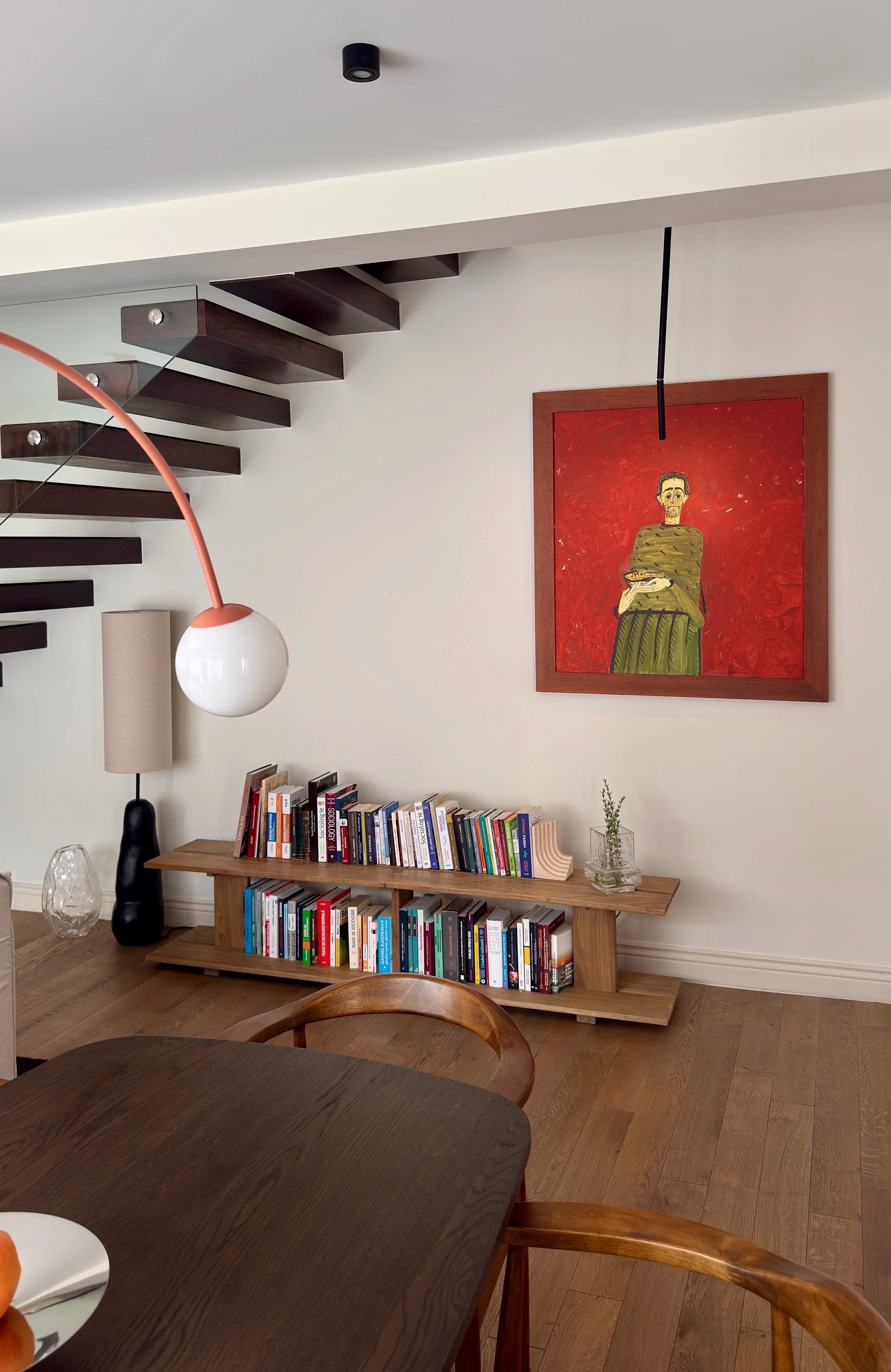 Interior of a modern living space with a wooden bookshelf, artwork of a woman on a red background, a floor lamp, and a staircase with dark wood steps and glass railing.