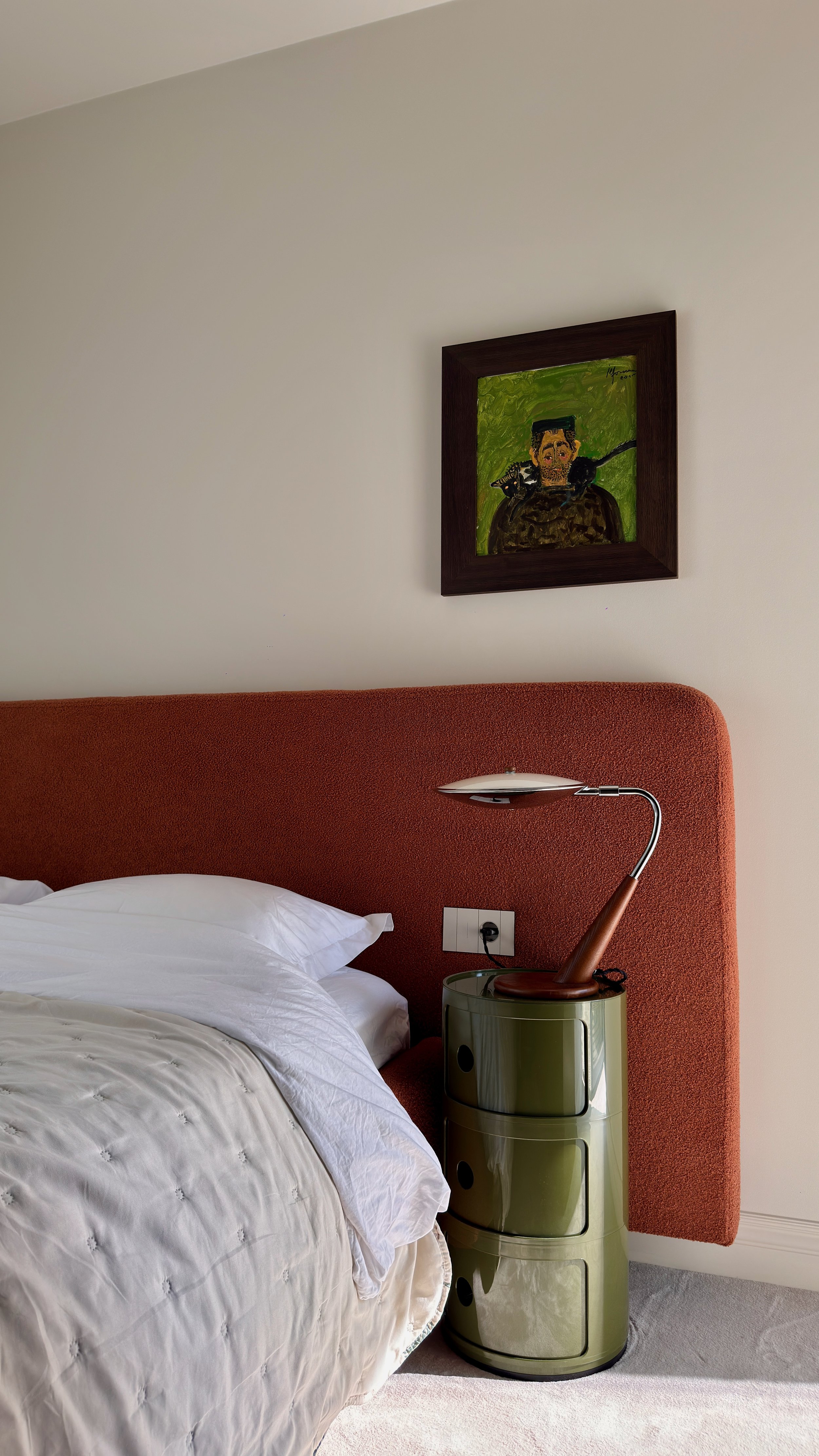 A bedroom with a bed, white bedding, an orange headboard, a green nightstand with drawers, a curved desk lamp, a framed painting of a man with two black cats on his shoulders, and a white wall.