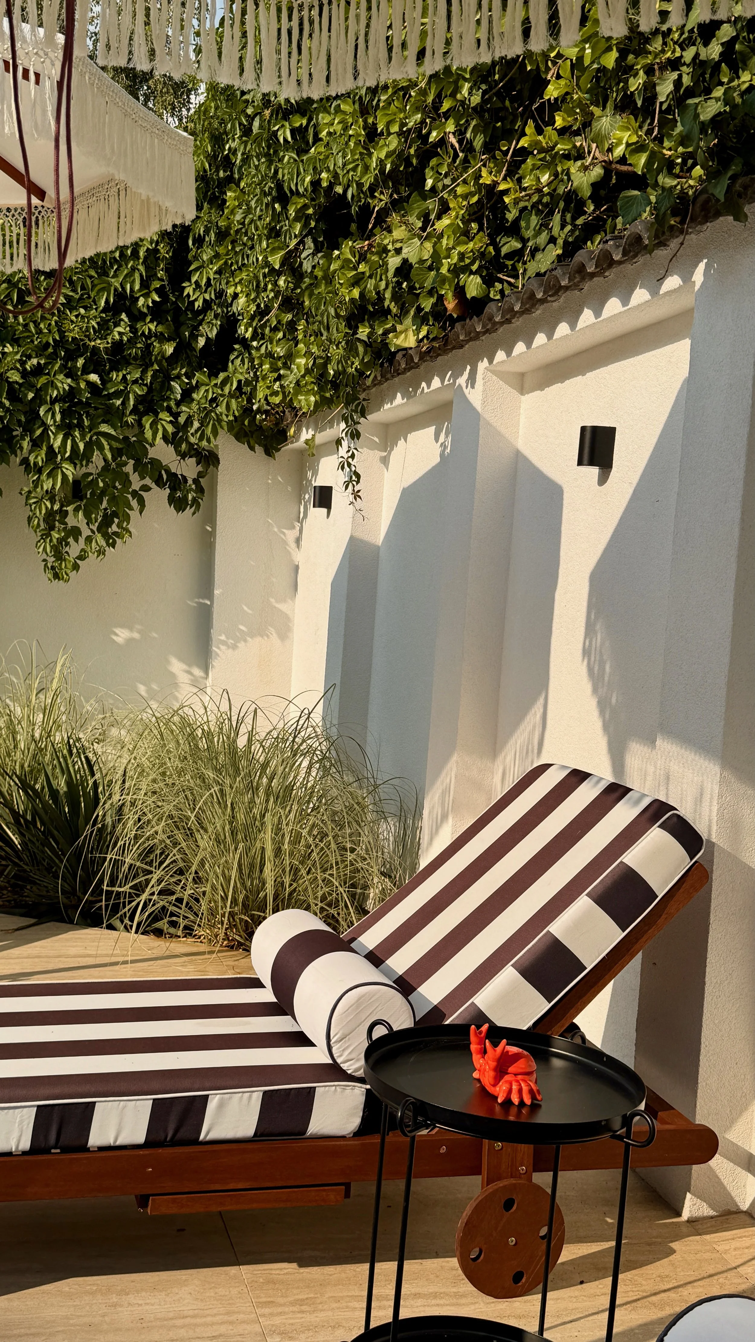A striped outdoor lounge chair with a cylindrical pillow and a small black side table with a red lobster-shaped decorative item, set against a white wall with wall-mounted lights, greenery, and a canopy overhead, part of a patio or garden area.