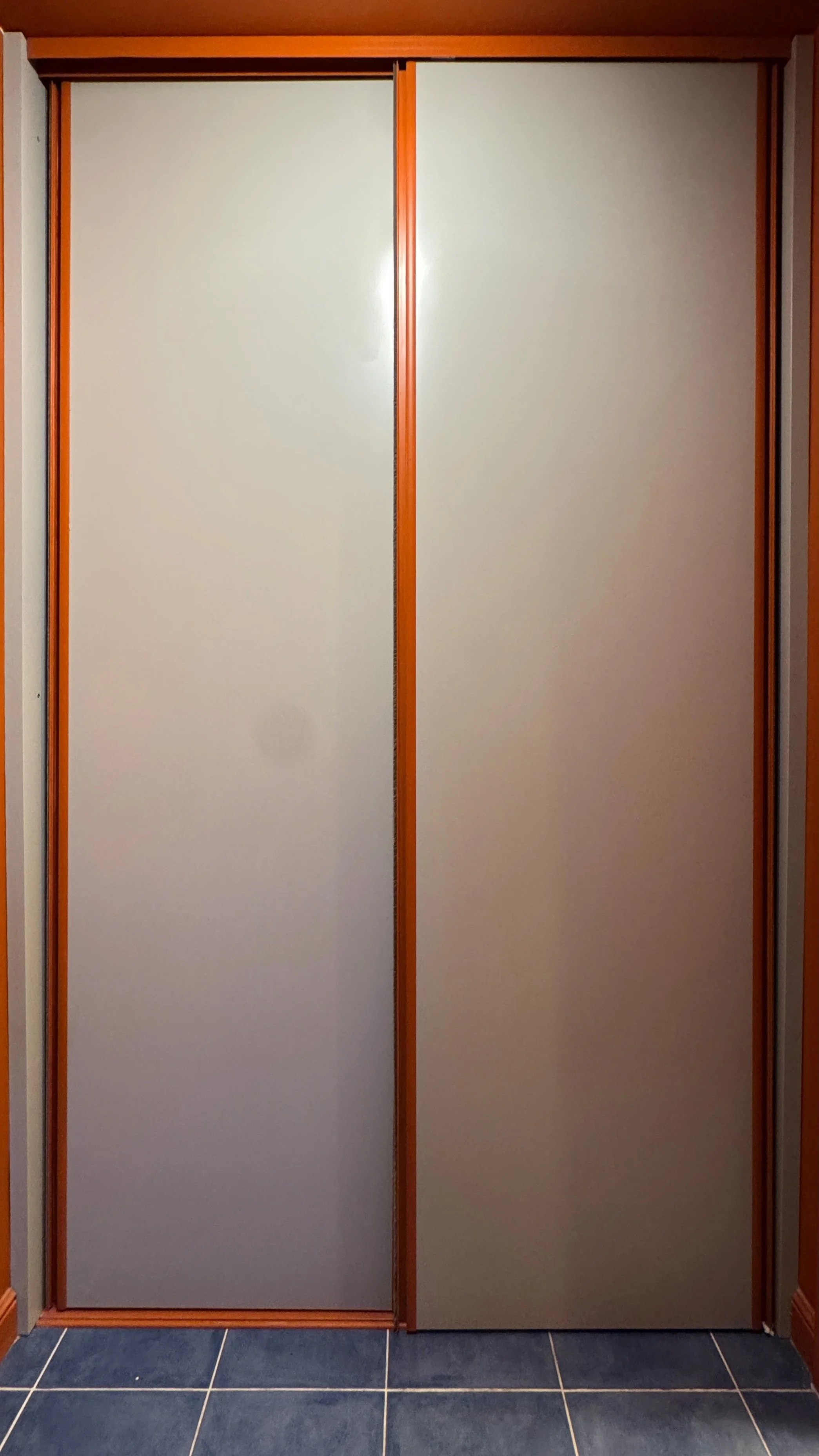 A closed sliding closet door with beige panels and orange-brown frames, set against a wall with dark blue tiled flooring.