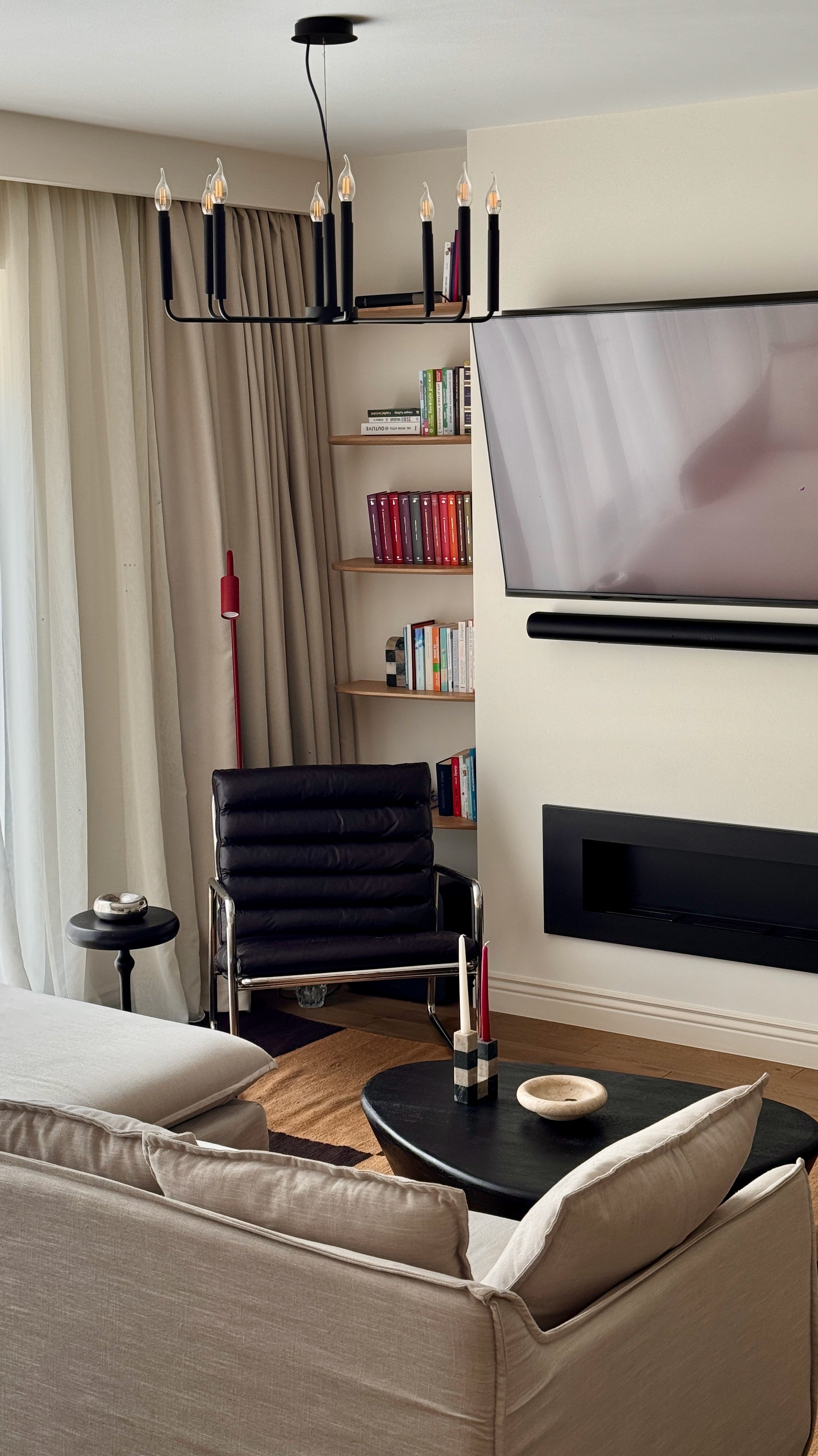 Living room with beige sofa, black leather chair, black coffee table, flat-screen TV, beige curtains, and wooden bookshelves.