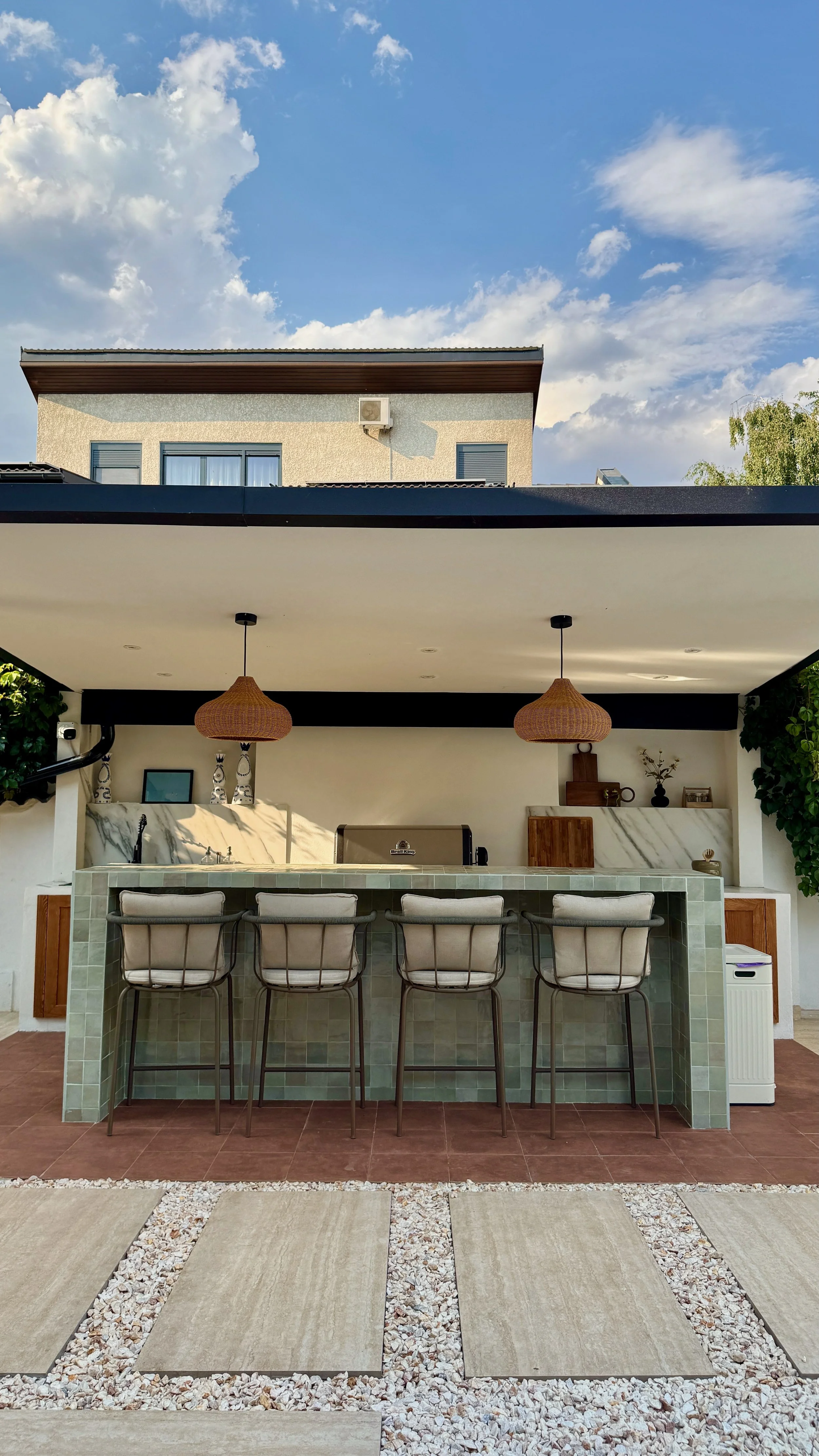 Outdoor kitchen with bar and bar stools, shaded by a roof, with string lights hanging from ceiling, and modern decor including potted plants, vases, and small sculptures, overlooking a paved patio and a two-story residential building.