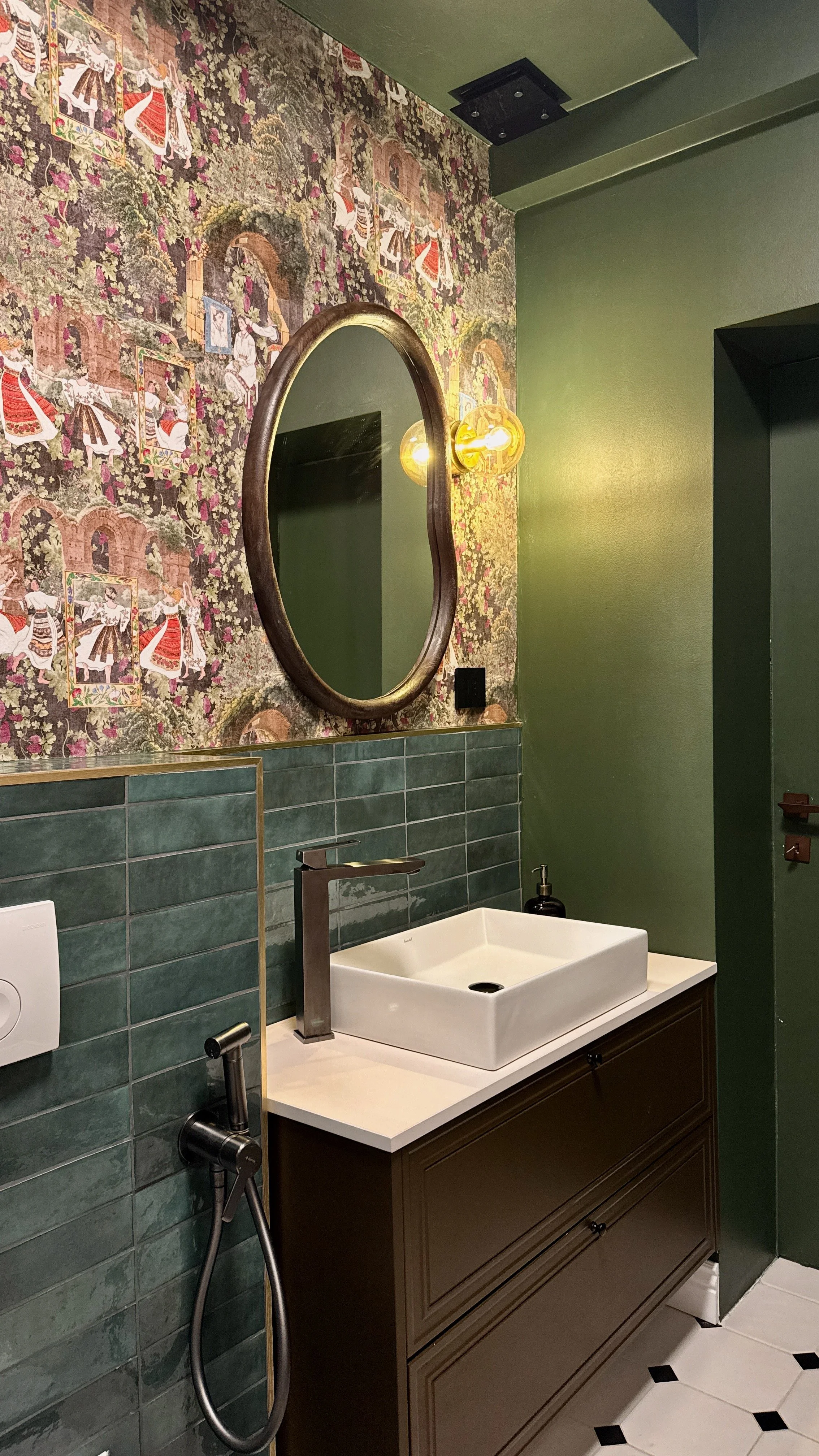 A bathroom featuring a green wall with floral and traditional patterned wallpaper, a wooden-framed oval mirror, contemporary brass light fixture, green tiled wall, a white modern sink, and a brown vanity cabinet.