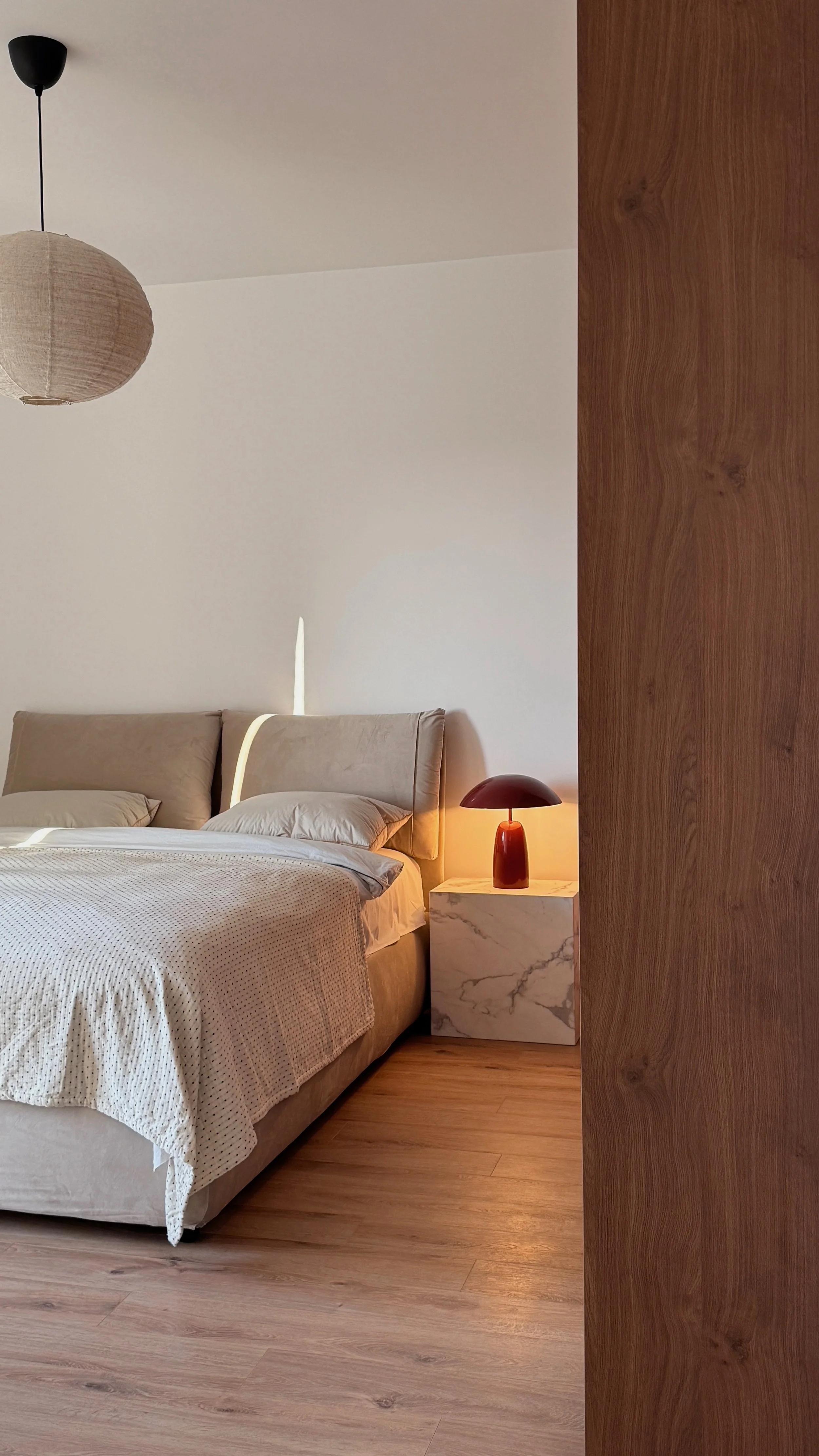 A bedroom corner with a bed, pillows, and a bedside table with a red lamp. The room has a wooden wall and light-colored flooring.