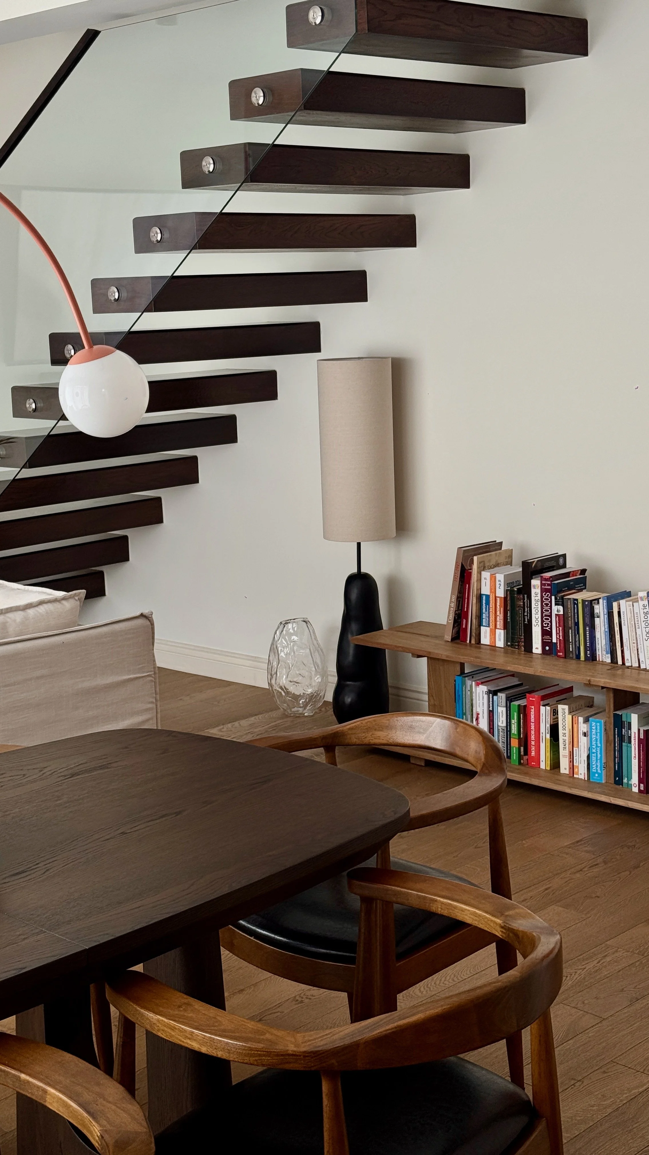 Living room with floating dark wood stairs, a modern black and beige floor lamp, a bookshelf filled with books, a glass vase, a beige couch, and a wooden dining table with chairs.