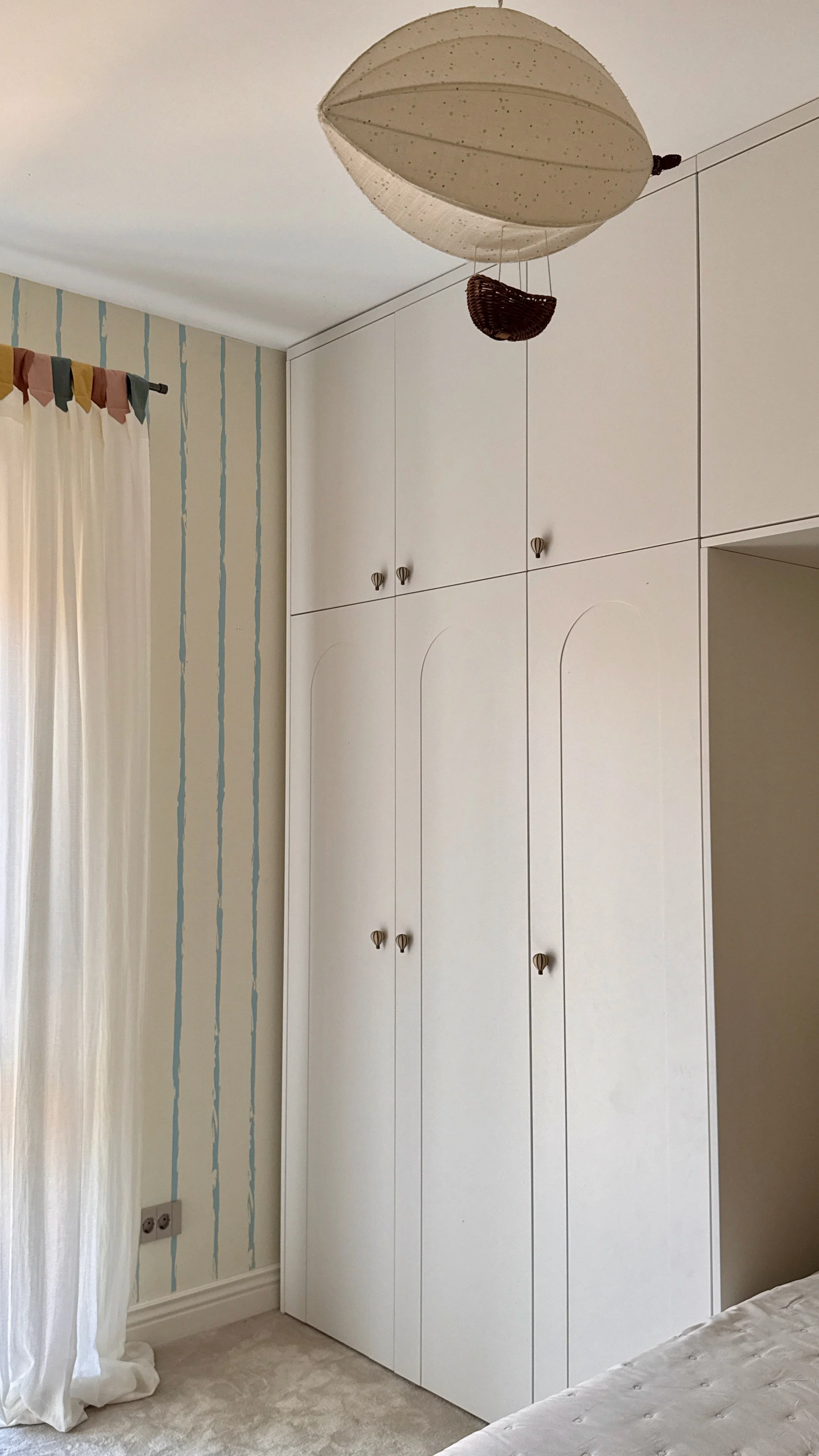 A white wardrobe with multiple doors is next to a light-colored wall with vertical blue decorative stripes. To the left, there is a white curtain hanging from a black curtain rod. The bottom of the wardrobe and the curtain are close to a cream-colore
