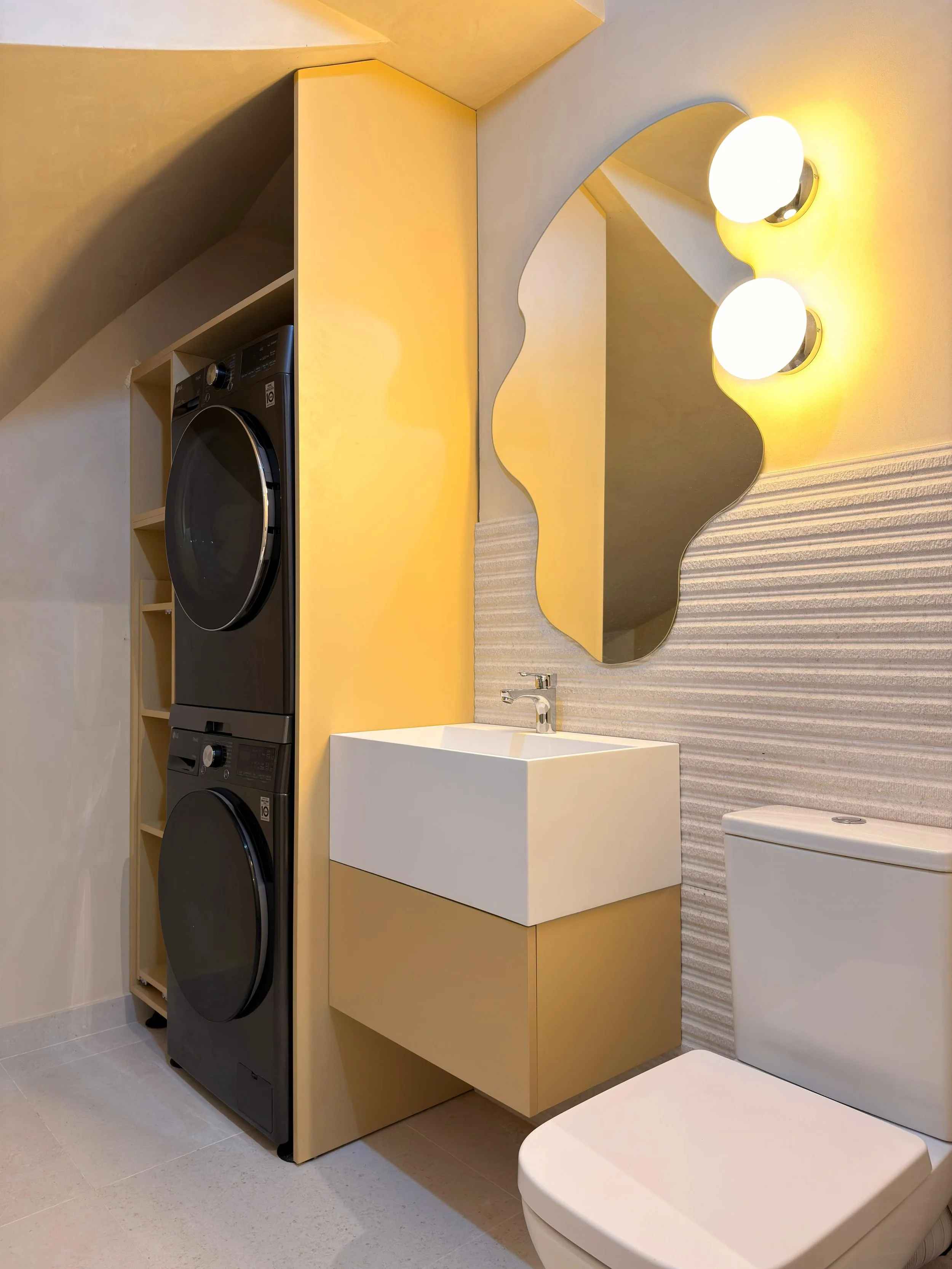 A modern bathroom featuring a stacked washer and dryer set in black, a yellow vanity with a white rectangular sink, a wavy-edged mirror, and two globe-style wall lights emitting warm yellow light.