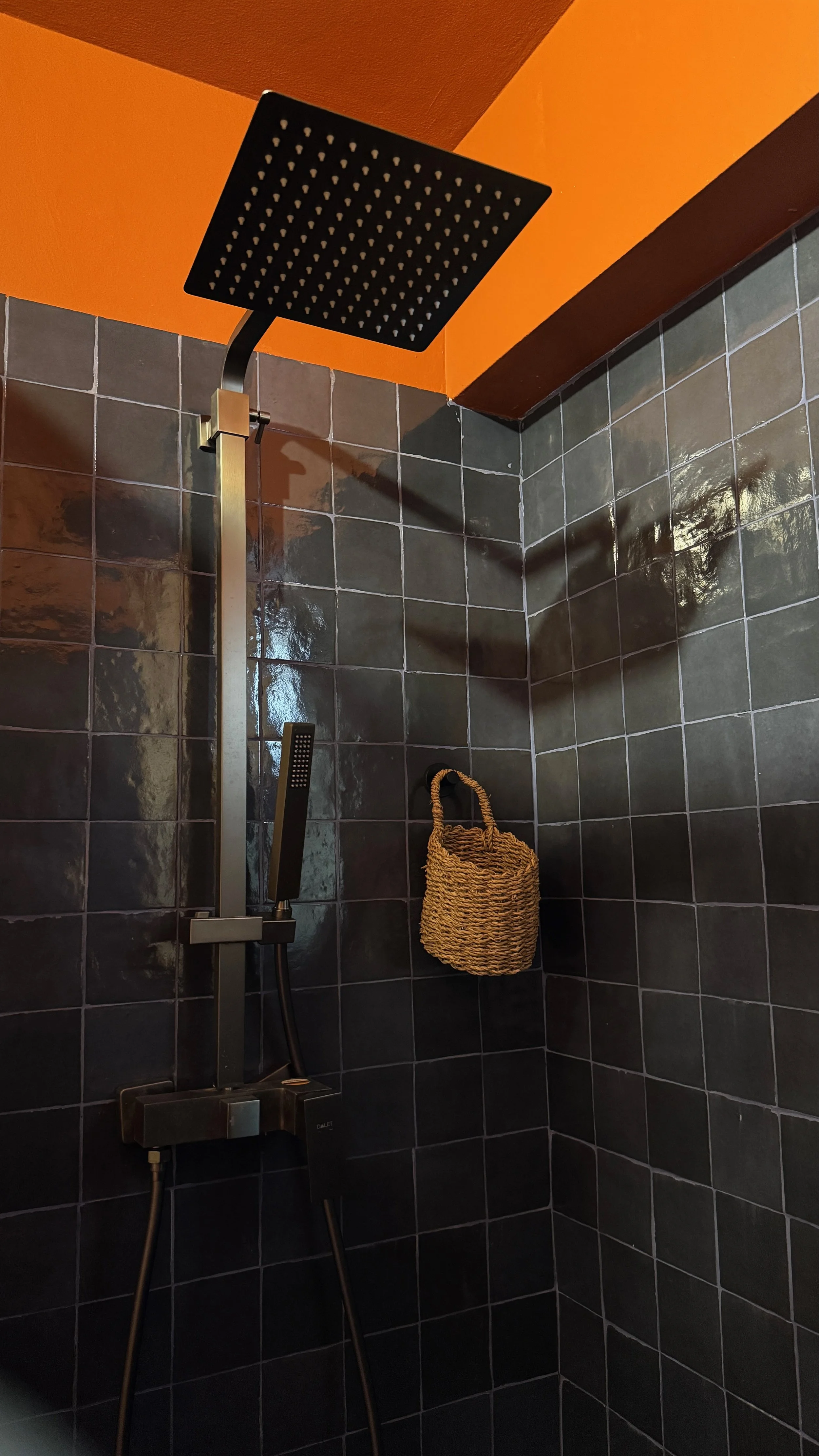 Shower with black tiles, orange ceiling, black overhead shower head, handheld shower, and a woven basket hanging on the wall.