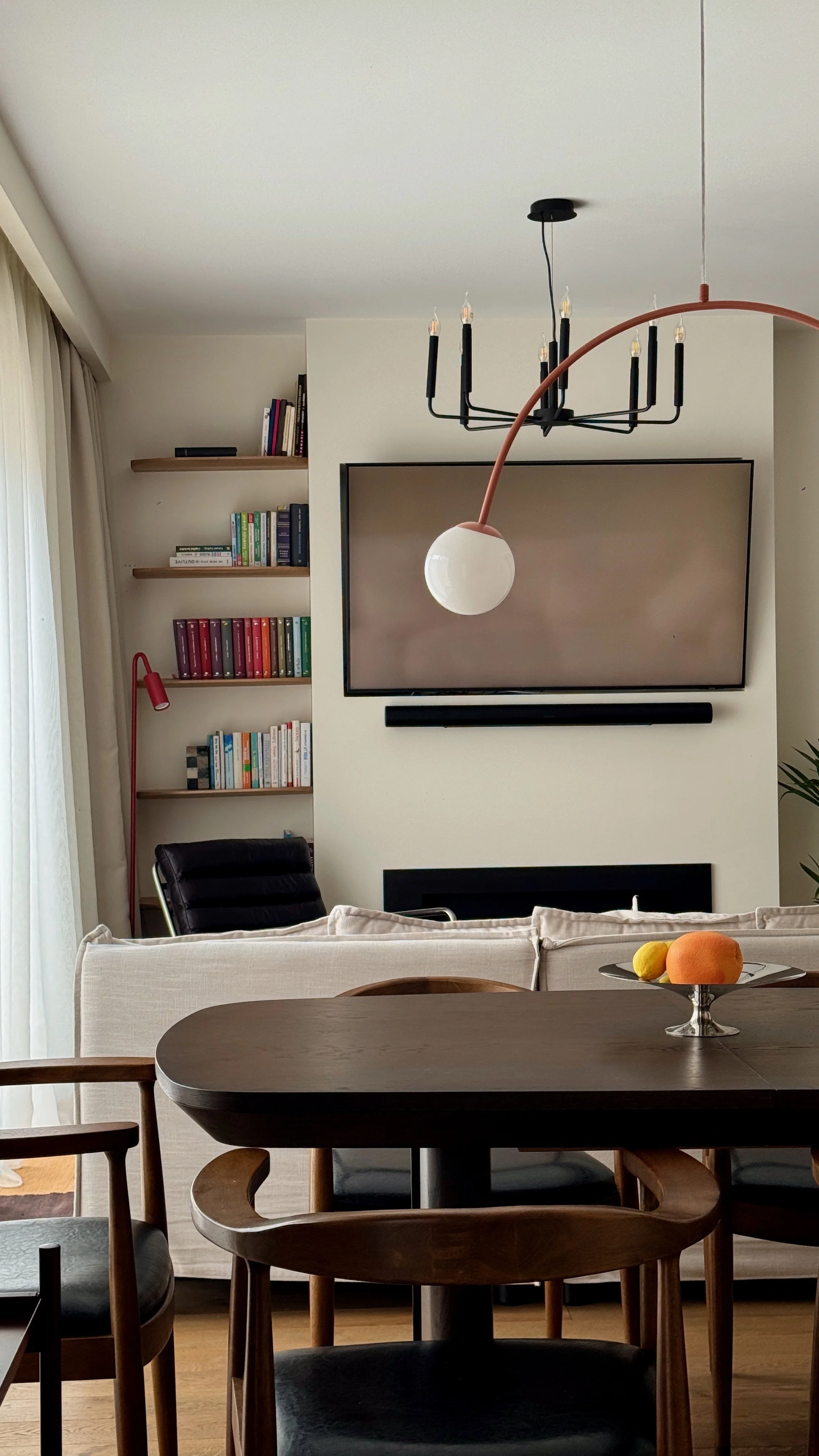A living room with a mounted flat-screen TV, a black soundbar, a black chair, a white sofa with pillows, a wooden dining table with chairs, a bowl of oranges and lemons, a black chandelier, and bookshelves with books and a red lamp.