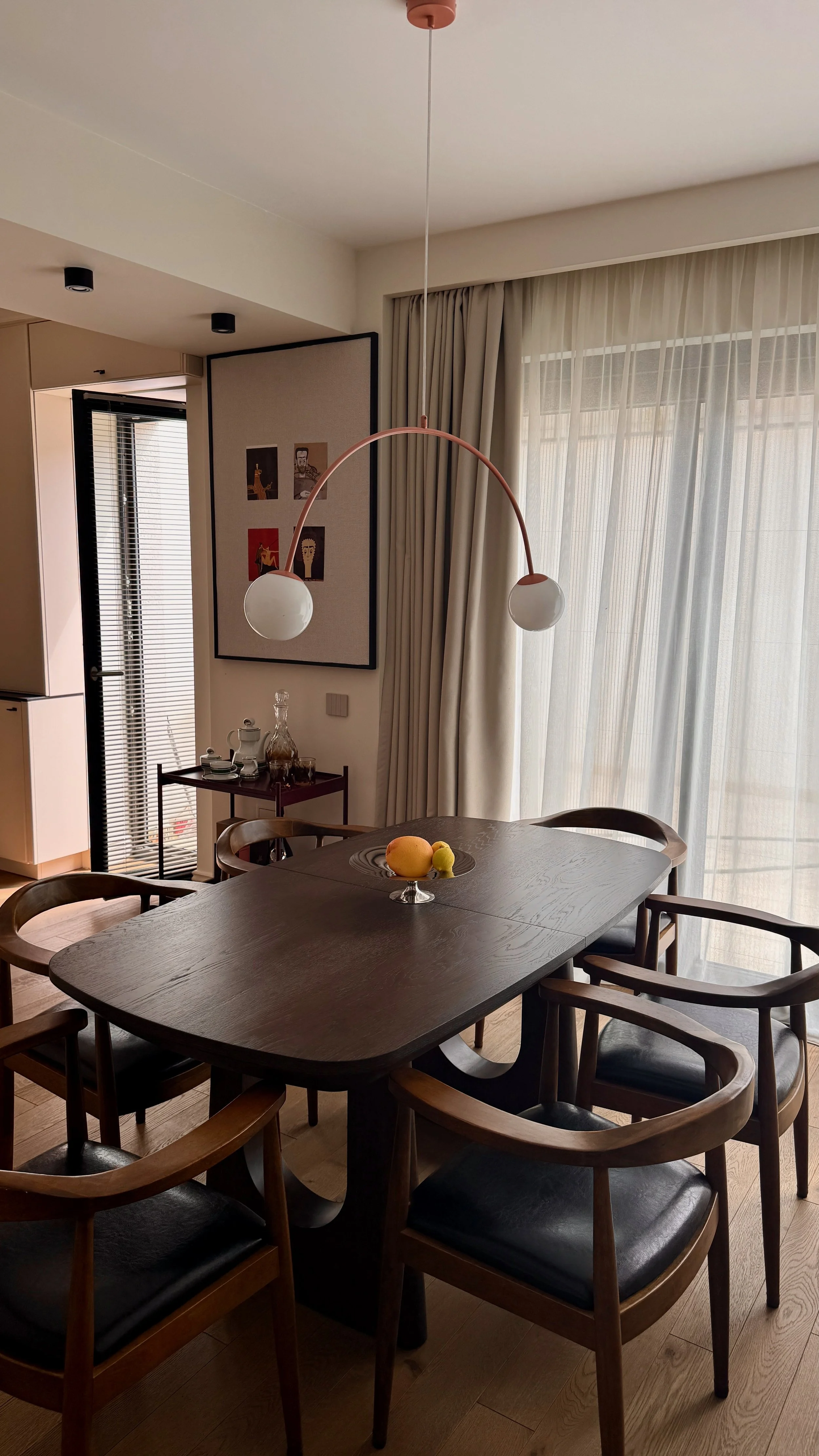 Dining room with wooden table and chairs, fruit bowl, large window with sheer curtains, modern hanging ceiling lamp, and wall art.