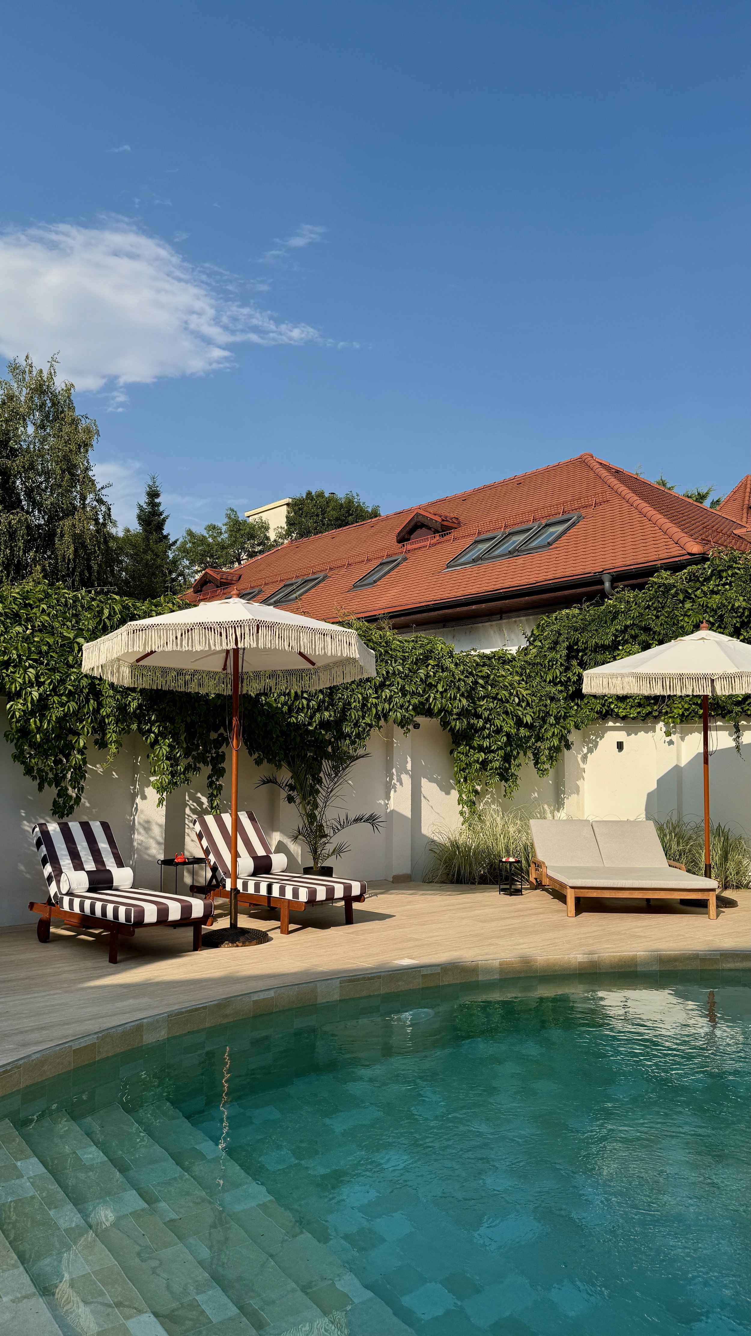Swimming pool with striped lounge chairs, umbrellas, and greenery, against a house with a red-tiled roof under a blue sky.